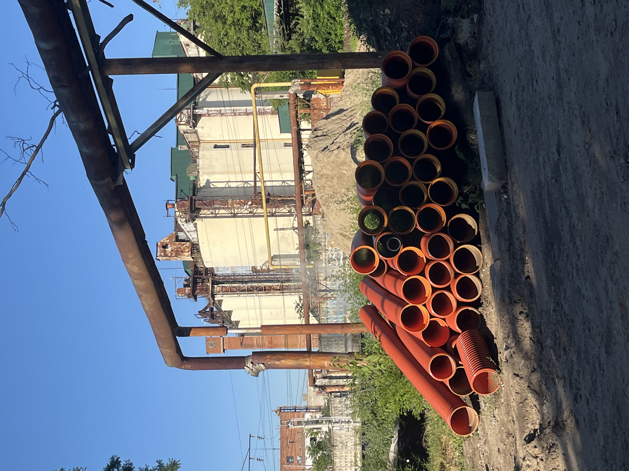 A pile of orange, plastic pipes lies on the ground in front of an industrial facility under a clear blue sky. The pipes, some smooth and others corrugated, are stacked and scattered. In the background, a large, light-colored industrial building with various metal structures and pipes is visible. Trees and green foliage are present to the right and left of the facility. The scene appears to be in an outdoor industrial area.
