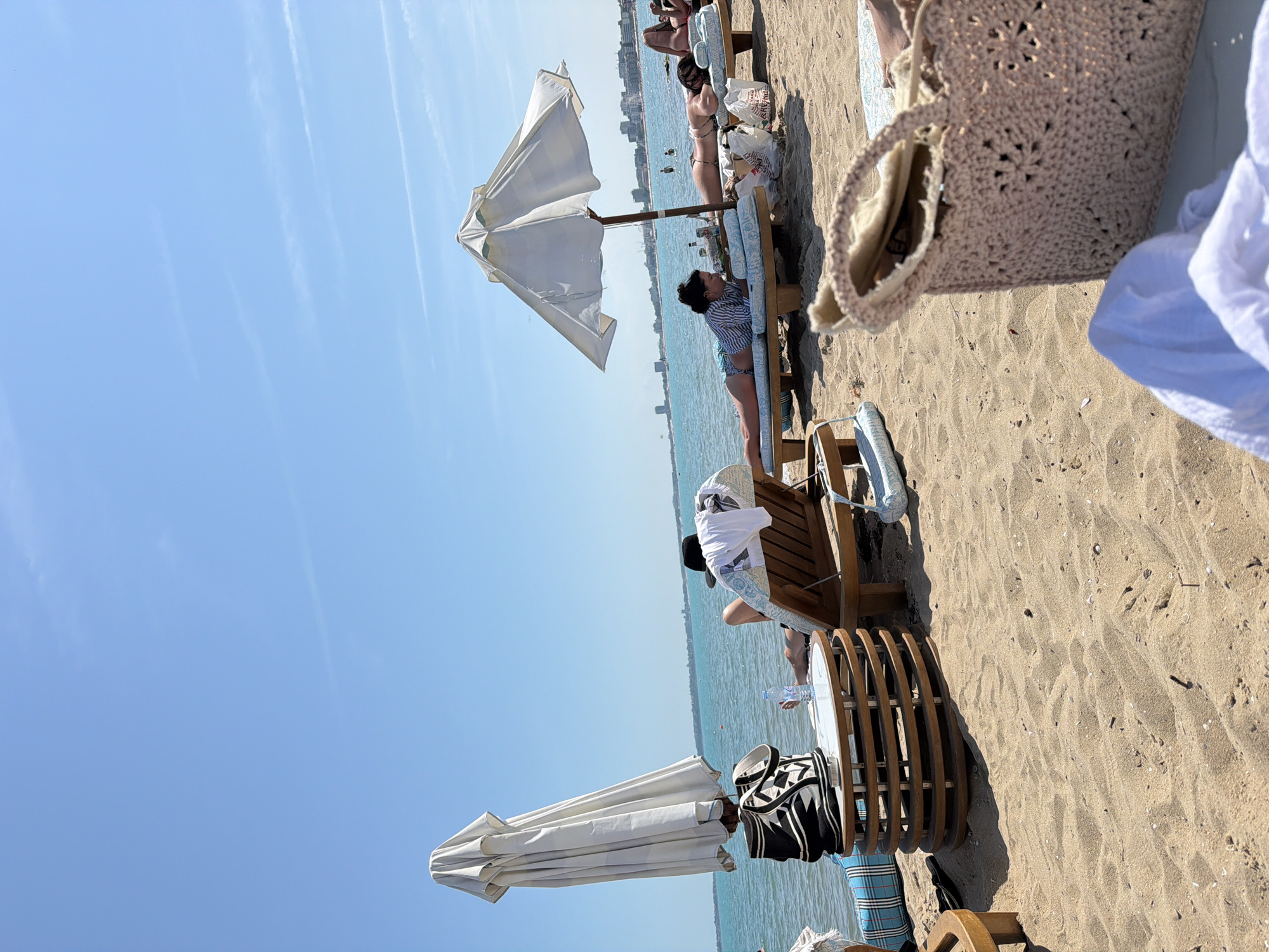 This image captures a sunny day at a sandy beach with a clear blue sky. The foreground shows light brown sand with scattered small shells and debris. In the immediate foreground, a light pink crocheted tote bag with star-shaped patterns is partially visible on the right, next to a white towel or piece of clothing.
In the midground, several beach items and people are arranged. On the left, a large, closed white beach umbrella stands upright, casting a shadow. Beside it is a circular wooden table with a light-colored top, on which a black and white patterned tote bag rests. A blue and white striped roll-up mat is partially visible next to the table. Further to the right, a person with dark hair, wearing a striped shirt and patterned swimwear, lies face down on a light blue cushioned lounge chair under an open white beach umbrella. They appear to be looking at a device held in their hands. A clear plastic water bottle is on the sand near their lounge chair.
To the right of the open umbrella, another person lies face down on a similar light blue cushioned lounge chair, sunbathing. Their light-colored swimwear is visible. Behind them, another individual is also sunbathing on a lounge chair, and a white plastic bag with red writing is on the sand near their feet. Further to the left, under the closed umbrella, a person's legs are visible on a lounge chair, next to which a black hat is placed.
The background features the calm, turquoise-blue sea, which extends to the horizon. Several buoys or small boats are scattered across the water. Along the distant horizon, a cityscape with various buildings is visible, suggesting a coastal town or city. The sky is bright blue with a few faint white contrails from airplanes. The overall scene depicts a relaxed beach environment on a clear, sunny day. The location is Unknown, Unknown.