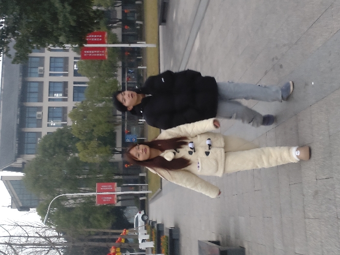 Two individuals are depicted walking outdoors on a paved pathway in the city of Jiangjin, China. The person positioned on the left is a female with long dark brown hair and eyeglasses, dressed in a cream-colored, fuzzy two-piece outfit consisting of a jacket with black toggle closures and matching pants, along with light brown slip-on footwear. She is looking slightly to her left. The person on the right is a female with short dark hair, wearing a black puffer jacket, light grey pants, and blue slip-on shoes. Her gaze is directed slightly upwards and to her right. Both individuals are walking from the left to the right side of the frame.
In the background, a multi-story building with a light-colored facade, multiple windows, and exterior air conditioning units is visible, partially obscured by various trees, some bare and some with green foliage. A section of the building features a dark, sloped roof. Several tall poles display red banners adorned with white Chinese characters. A white SUV is partially visible parked on the far left, behind a low planter or barrier. The ground consists of a walkway paved with rectangular grey stones. A white, curved streetlight pole is positioned on the left side of the frame.