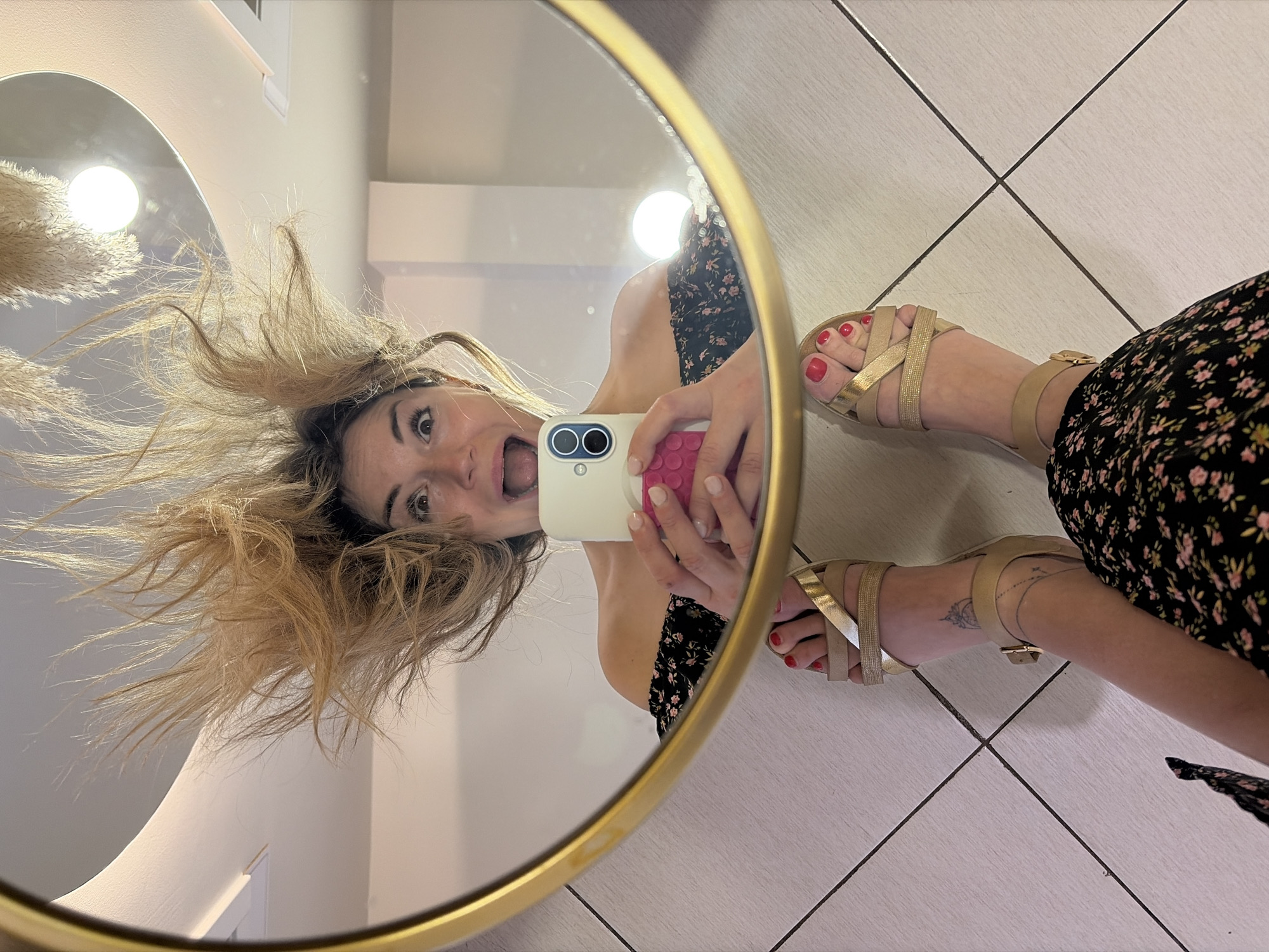 A woman takes a selfie in a mirror. The mirror is round with a gold frame. The woman has blonde hair and is wearing a floral dress and gold sandals. She is standing on a tiled floor. The location is most likely an interior space in an urban setting, potentially in Budva, Montenegro.