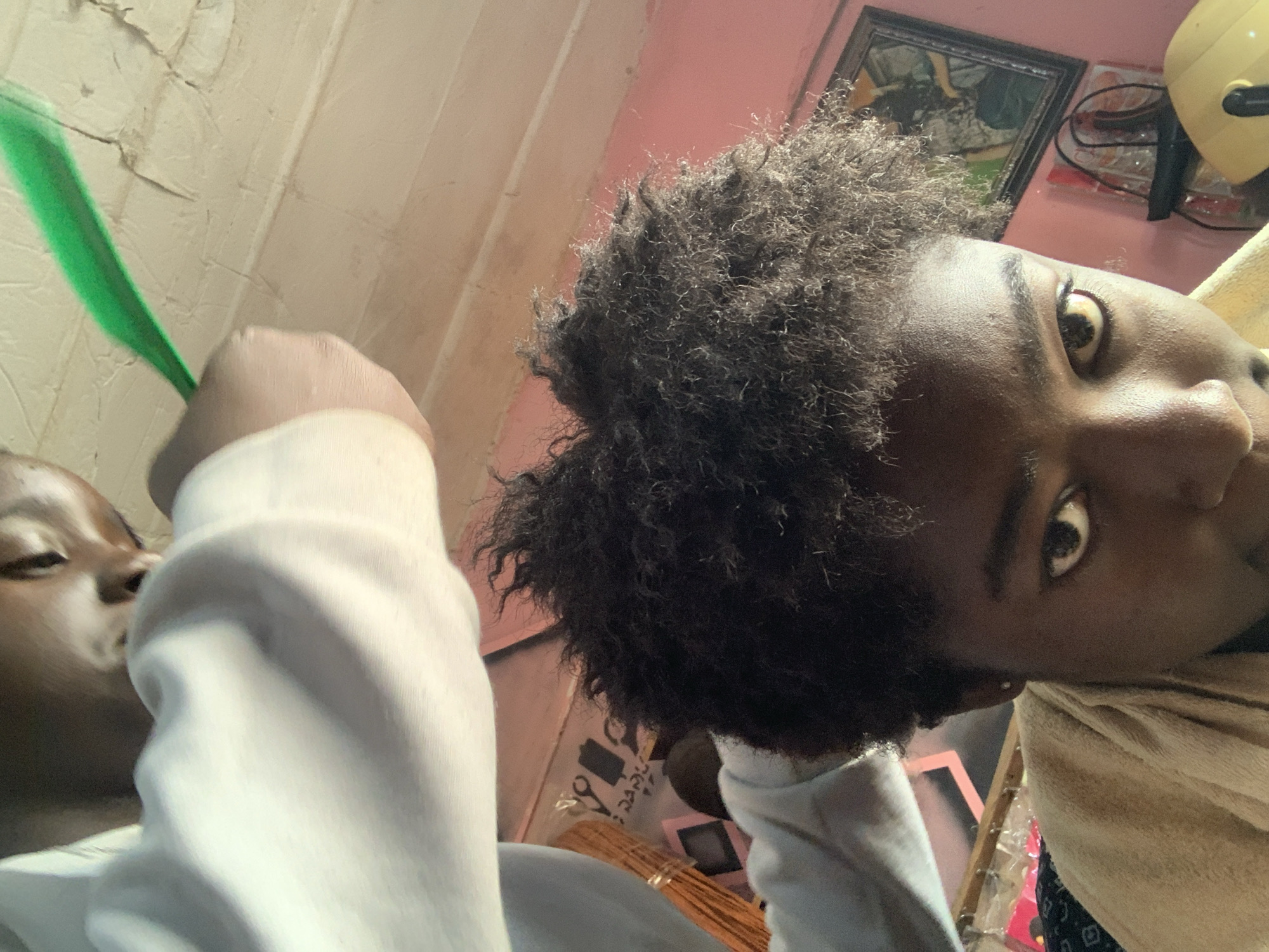A close-up, slightly distorted photo shows two Black individuals indoors. One person is holding a green object, possibly a pen or wand, near the face of the other. The environment contains domestic elements like pink walls, framed artwork, and possibly hair styling equipment. The location is potentially Rafin Par, Nigeria.