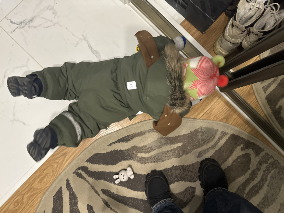 A baby in a green snowsuit lies on a floor. They are wearing a red and green striped hat. Nearby, a small white toy bunny sits on a zebra-striped rug. The lower portion of a person with blue jeans and black shoes is visible at the bottom edge of the rug. Behind the child is a mirrored closet with a pair of white FILA sneakers on the top shelf. The floor is a mixture of tile and wood.