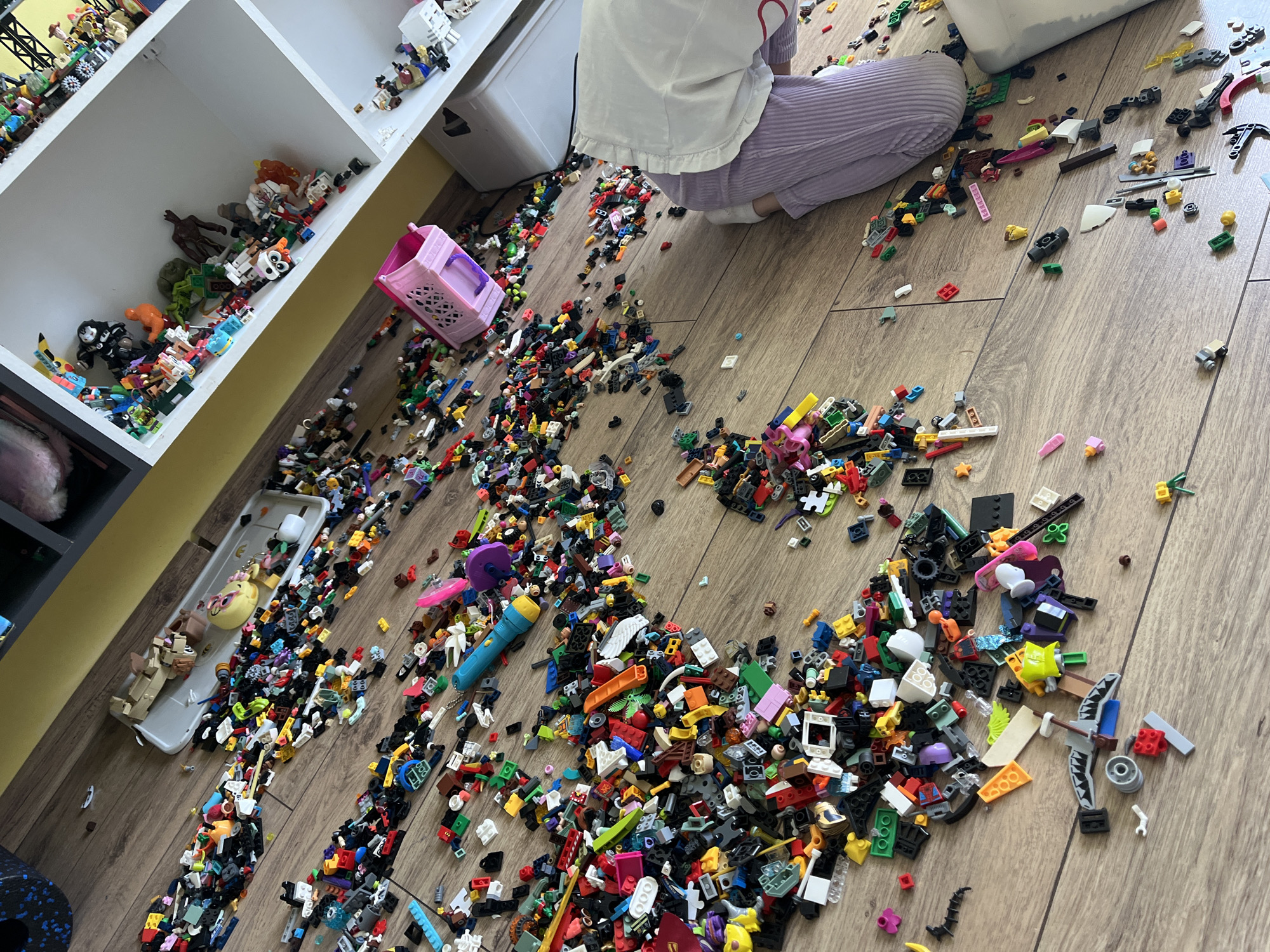 The image depicts a child sitting on a wooden floor surrounded by a large number of Lego bricks. The child is wearing a white shirt and purple striped pants. To the left of the child is a white shelf unit filled with various toys and figurines. A pink plastic basket is also near the shelf. The floor is covered with a dense layer of Lego bricks in a variety of colors and sizes. A white storage container is located behind the child.