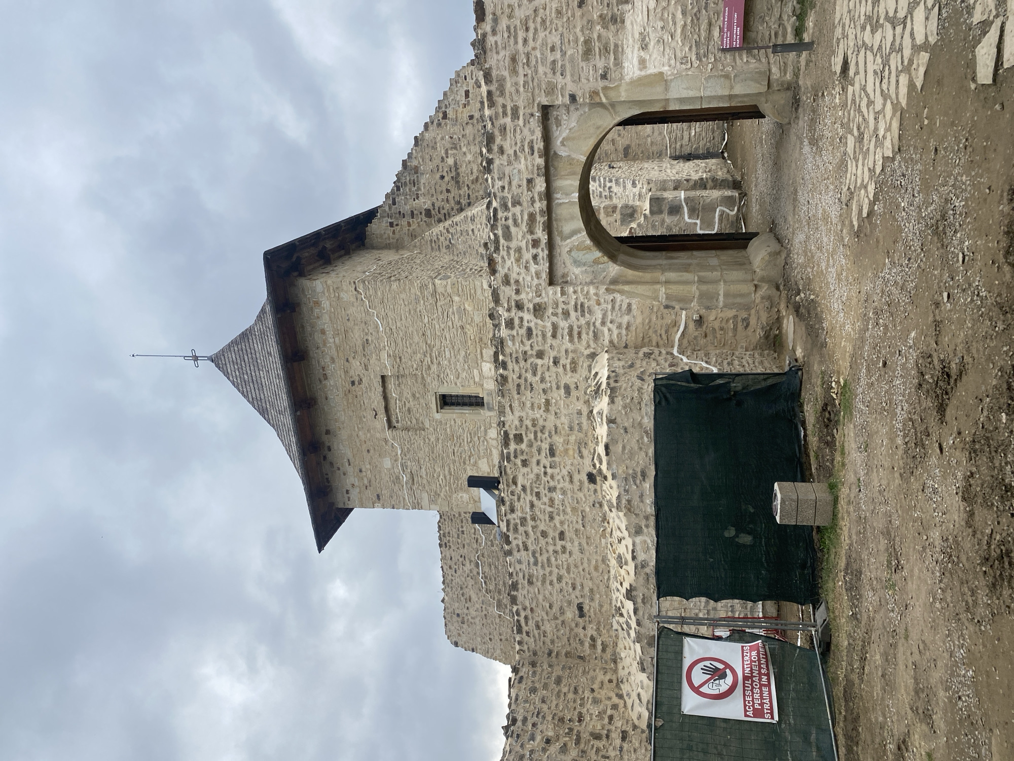 The image shows the fortress of Stephen the Great in Suceava, Romania. The fortress is made of stone and has a large tower with a wooden roof. There is an arched entrance to the fortress. In the foreground, there is a construction fence with a sign that says "ACCESS PROHIBITED TO UNAUTHORIZED PERSONS ON THE CONSTRUCTION SITE." The sky is cloudy.
