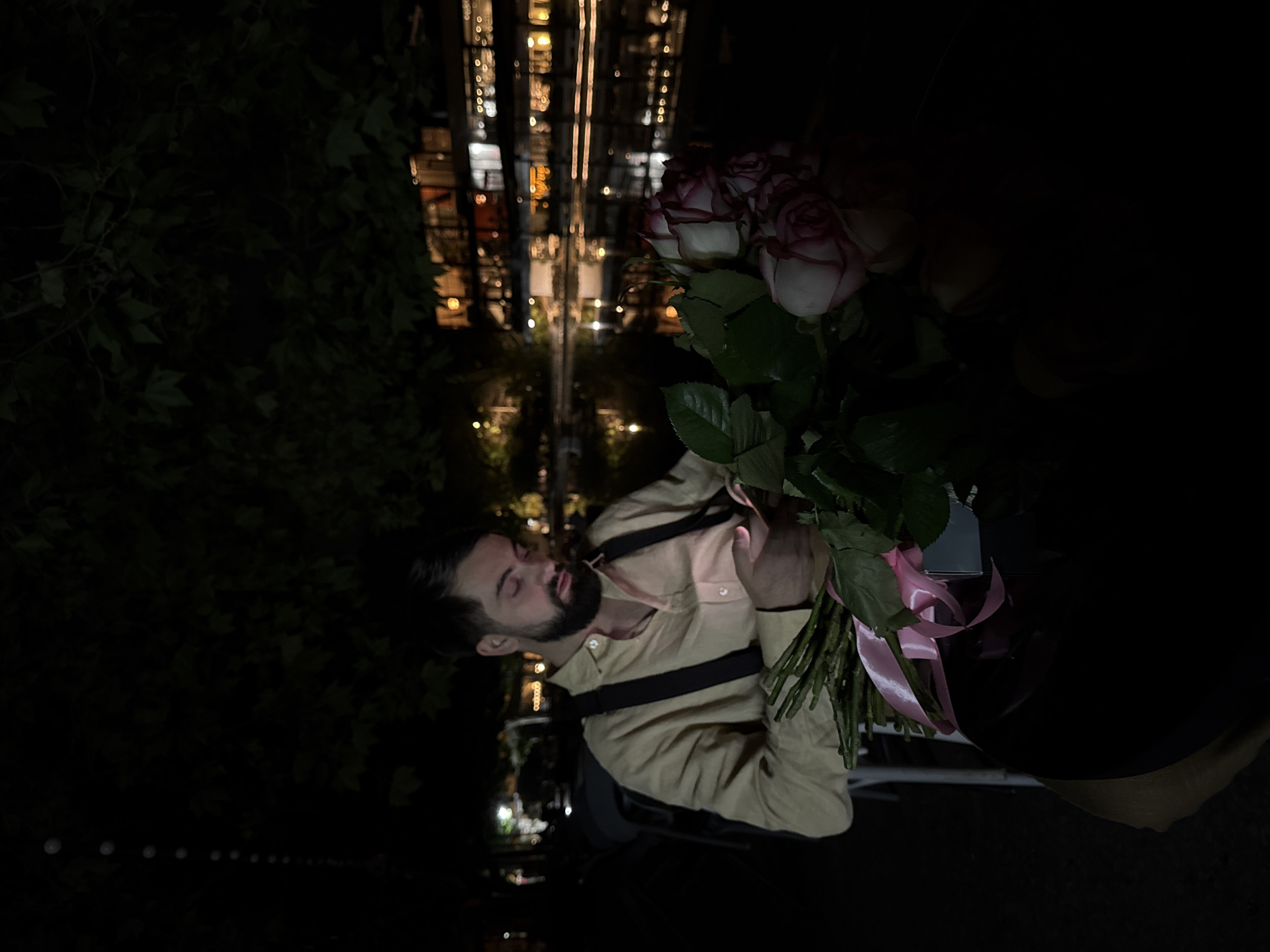 The image, rotated 90 degrees clockwise, captures a night scene featuring a man holding a bouquet of roses outdoors. The man, with a beard and dark hair, is positioned in the lower center. He wears a light-colored (possibly beige or tan) collared shirt with two dark, horizontal straps across his chest, suggesting suspenders or part of an accessory. His eyes appear closed or are cast downwards, and his mouth is slightly open, conveying a pensive, emotional, or perhaps sleeping state.
He holds a large bouquet of roses in his left arm, consisting of white roses with prominent pink or reddish edges, tied with a bright pink ribbon. The roses are held close to his chest and partially obscure the right side of his body.
The background is dark, indicating nighttime. To the left, there is dense, dark foliage, likely bushes or trees. To the right and behind the man, blurred city lights create an out-of-focus backdrop, suggesting buildings or a cityscape. The reflections of these lights are visible below, indicating a wet surface or water, possibly a canal or large puddle. The overall lighting is dim, with the man and flowers being the most illuminated subjects, likely by an artificial light source close to the camera.
No text is visible in the image. The scene is set in Yerevan, Armenia, at night, suggesting a quiet or intimate moment in an urban environment.