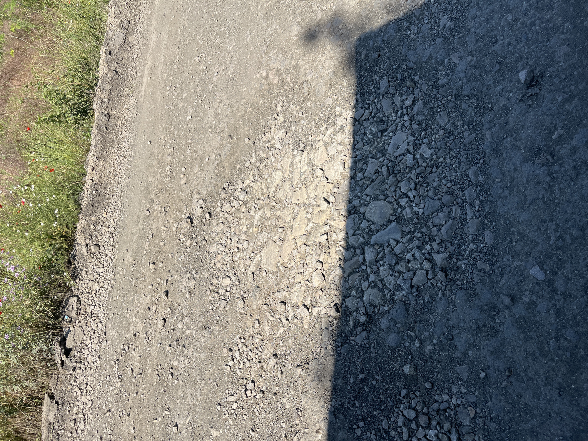 The image shows a rough, unpaved road or path in the city of Poroy, Bulgaria. The ground is covered with gravel and stones of various sizes. In the upper part of the image, there is a patch of dry grass and wildflowers, including some red poppies and white flowers, growing along the edge of the path. The lower part of the image is cast in shadow, revealing a darker, more compacted surface with scattered stones. The shadow suggests the presence of an object or person outside the frame, casting a silhouette of a head and shoulders. The overall scene depicts an outdoor, somewhat unkempt environment.