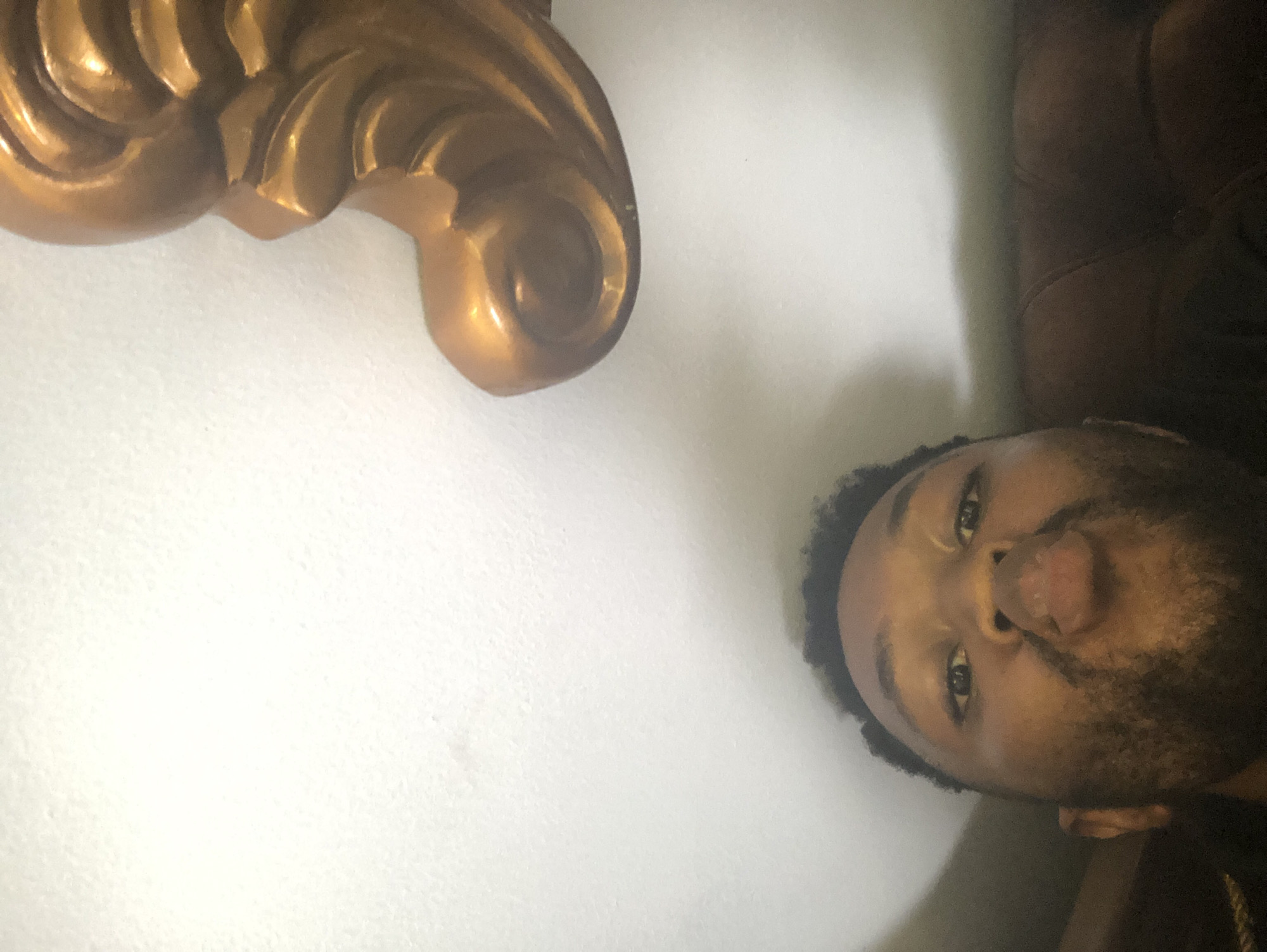 The image shows a person with dark hair and a beard, lying down and looking up at the camera. Their face is close to the lens, with their mouth slightly open. They appear to be resting on a brown, textured surface, possibly a leather couch or chair. In the upper left corner of the image is a gold-colored, ornate architectural detail, possibly a piece of molding or a decorative element attached to a wall. The wall behind the person and the gold detail is plain and light-colored, likely white. The overall lighting of the scene is soft and indirect. No location details are available.