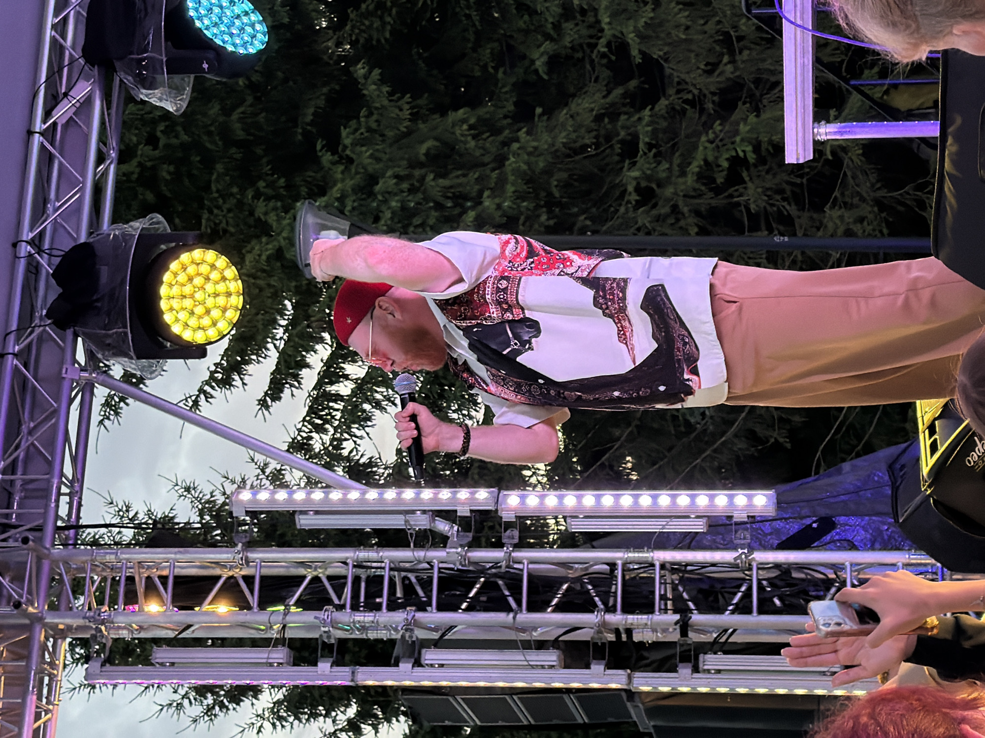An outdoor evening concert stage is illuminated by purple and yellow lights. A man wearing a red beanie, glasses, and a patterned shirt is singing into a microphone. He is holding a plastic cup in his raised right hand. The stage is adorned with vertical light strips and speakers. In the foreground, audience members are visible, some holding up phones to capture the performance. Trees form the backdrop against a dusky sky.