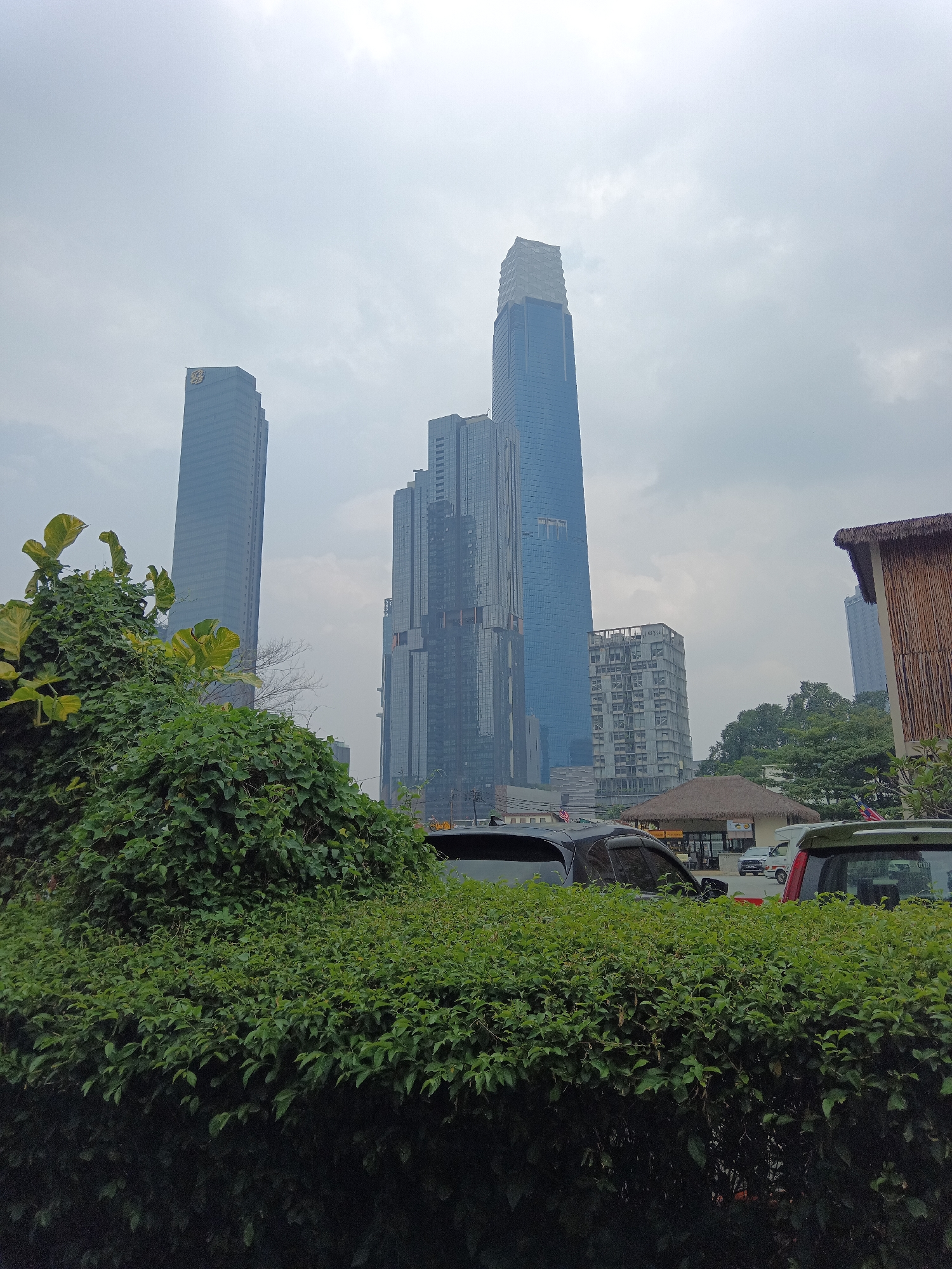 The image depicts the skyline of Kuala Lumpur, Malaysia, featuring several modern skyscrapers. In the foreground, lush green bushes and foliage dominate the lower half of the frame, with some vines trailing over a wall on the left. Behind the greenery, cars are parked or driving, including a dark SUV on the left and a lighter colored SUV on the right. A small building with a thatched roof is visible in the middle ground, appearing to be a cafe or restaurant.
Prominent skyscrapers pierce the overcast sky. The tallest and most striking is the blue glass skyscraper with a distinctive tapered top, likely the Merdeka 118, which is partially obscured by clouds. To its left, another tall, slender blue glass skyscraper stands, and further left is a wider, greyish-blue skyscraper. Several other buildings of varying heights and architectural styles are visible in the background, interspersed with trees and the hazy sky. The overall scene suggests an urban environment where modern architecture coexists with natural elements.