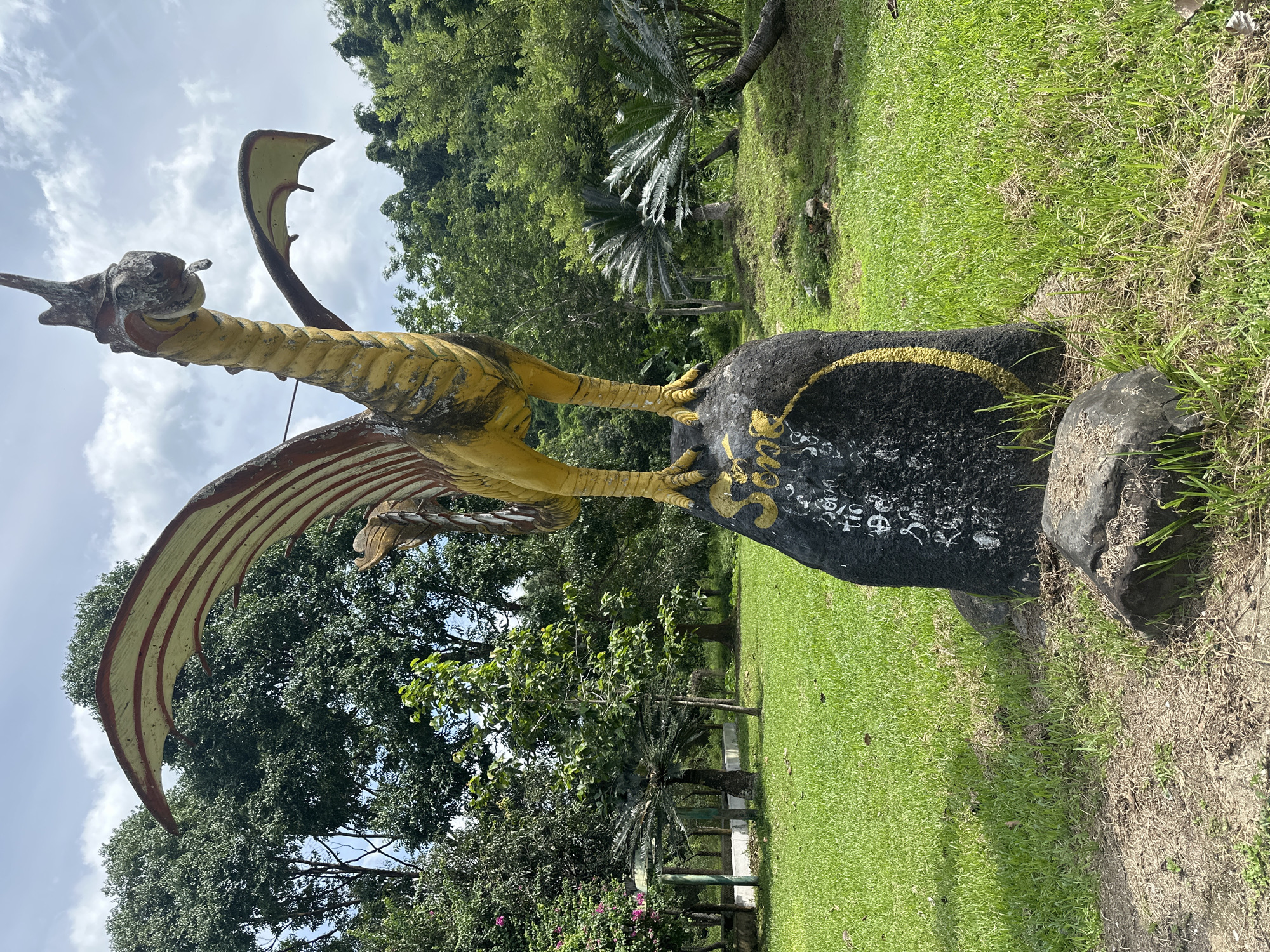 A weathered, yellow and red statue of a mythical creature, possibly a dragon or phoenix, stands prominently on a dark, roughly hewn rock in a grassy park. The statue depicts a creature with wings spread, a long neck, and a distinctive head with a pointed crest. The rock it stands on is inscribed with white lettering and a yellow circle. The surrounding environment is lush and green, with a well-maintained lawn, various trees, and cycad plants. In the background, a gentle slope covered in dense foliage suggests a natural, hilly landscape. The sky above is partly cloudy, with patches of blue visible. The scene appears to be an outdoor recreational area, possibly a park or garden, located in Khanh Vinh, Vietnam, given the context. There are no people visible in the image. The statue's aged appearance and the natural setting imply a tranquil and perhaps historical or cultural site.