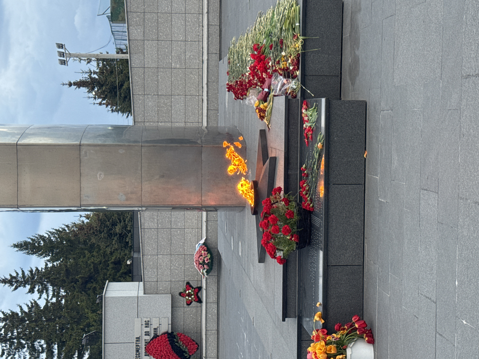 The image shows a war memorial complex located in Kazan, Russia. A large, silver-colored obelisk stands next to a black granite eternal flame with a five-pointed star design. Flames rise from the center of the star. Flowers, mostly red, are arranged on the steps and around the base of the memorial. Wreaths are placed at the base of a nearby plaque. The memorial is set on a grey, tiled plaza. Trees and buildings are visible in the background. The sky is overcast.