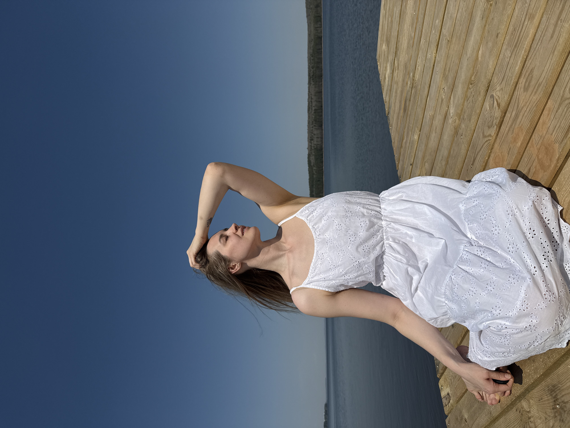 A woman in a white dress stands on a wooden dock, facing away from the camera. The dock is located in Chaikovsky, Russia, and the background consists of the Kama River and a clear blue sky. The woman has her right arm raised, touching her hair.