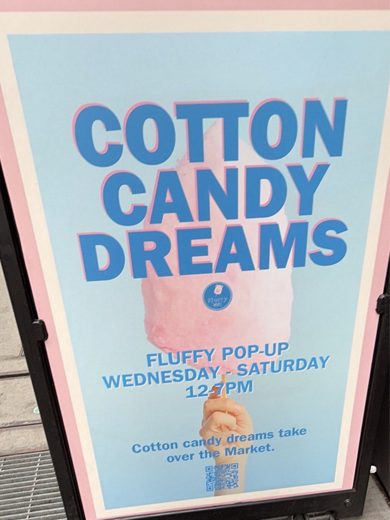 Here is a description of the image:
The image shows an advertisement sign for "Cotton Candy Dreams," a "Fluffy Pop-Up" shop. The sign has a light blue background with the words "Cotton Candy Dreams" in large, blue letters with a pink outline. Below that is a pink cotton candy held by a hand, along with the shop's logo. The words "Fluffy Pop-Up" and "Wednesday - Saturday" "12-7 PM" are written beneath the cotton candy, with "Cotton candy dreams take over the Market" underneath, followed by a QR code. The sign is framed with a black border and a pink outline. The image was likely taken outdoors in New York City, United States, since the shop is advertised as "taking over the market."