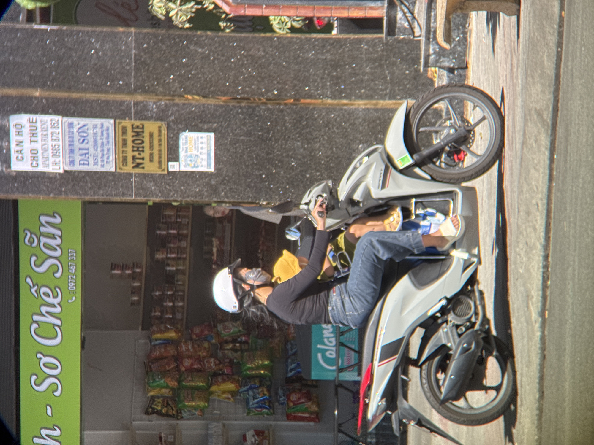 A woman wearing a black long-sleeved shirt, blue jeans, and a white helmet rides a silver and white scooter on a paved road in Nha Trang, Vietnam. She is wearing a mask and her hair is blowing in the wind. Behind her, a child wearing a yellow shirt and a yellow hat is sitting on the scooter. The scooter has red and black accents. To the left of the scooter is a shop with shelves filled with snacks and drinks. A sign above the shop entrance reads "h - So Che San" and a phone number. To the right of the scooter, a dark, textured wall has several signs attached to it. One sign is white and reads "CAN HO CHO THUE APARTMEN FOR RENT LH: 0985 872 852". Below that, another sign is light blue and reads "DAI SON". Further down, a yellow sign reads "CONG TY TNHH TMOH NT-HOME". A small, white sign with a blue border is attached below the yellow sign. In the background, a brick wall and some foliage are visible. The sun casts a shadow of the scooter on the ground.
