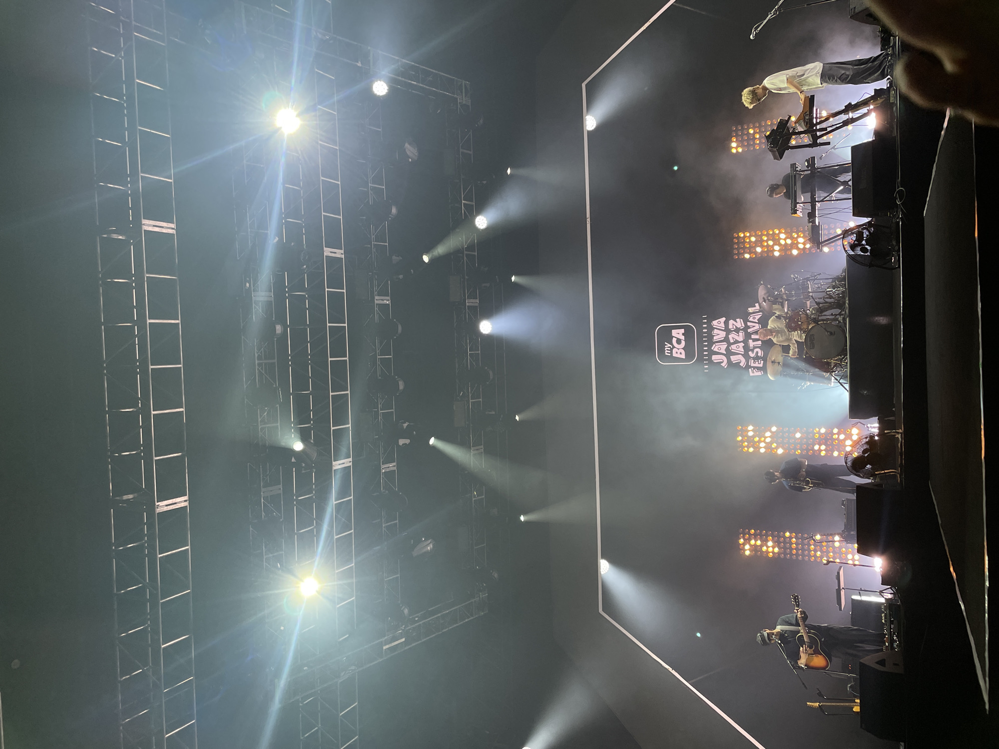 The image shows a stage at the International BCA Java Jazz Festival in Salembaran, Indonesia. A band is performing, featuring musicians on keyboards, drums, guitar, and other instruments. The stage setup includes a lighting grid above and rows of lights behind the performers. The stage is illuminated with spotlights.