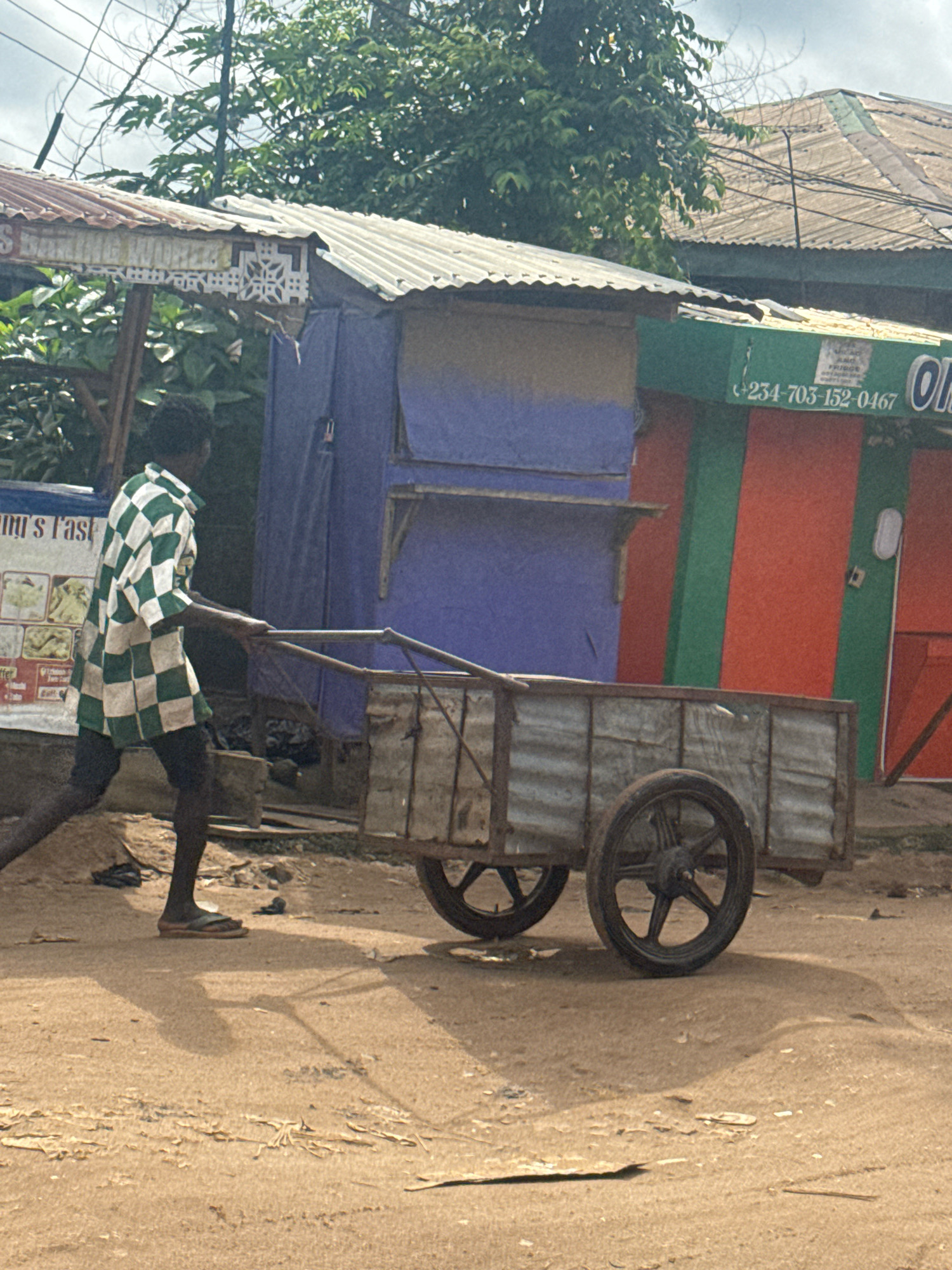 A man wearing a green and white checkered shirt and dark trousers is pushing a two-wheeled metal cart down a dirt road in Ihiagwa, Nigeria. The cart is laden with what appears to be goods or materials. Behind the man and the cart, there are several structures with corrugated metal roofs, one painted in shades of blue, red, and green, and another with a faded sign. Trees and vegetation are visible in the background, along with power lines and a cloudy sky. The ground is dusty and uneven. The scene suggests a commercial or residential area in a Nigerian city.