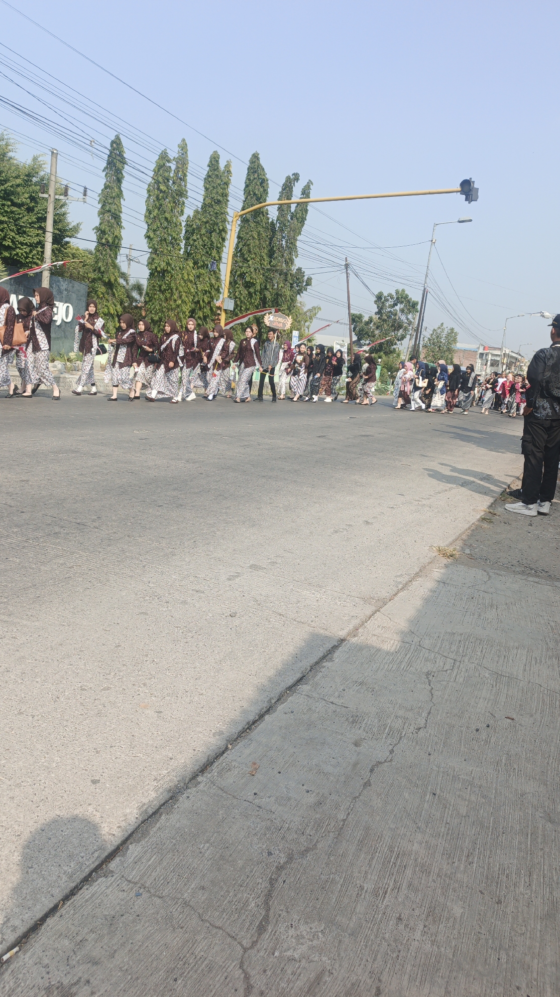 A group of people, many of whom appear to be students in traditional Indonesian attire, are participating in a procession on a street in Sumengko, Indonesia. The individuals on the left side of the frame are wearing matching brown and white patterned outfits with headscarves. They are walking in a line, some carrying bags. Further down the street, more people are visible, some in similar traditional clothing and others in more casual wear. A traffic light hangs over the road, with tall, slender trees lining the background. Power lines are strung across the sky. A person in a black shirt and pants, with white shoes, stands on the right side of the road, observing the procession. The road surface is paved with concrete, showing some cracks and wear. The lighting suggests it is daytime, likely morning or late afternoon, with clear skies.