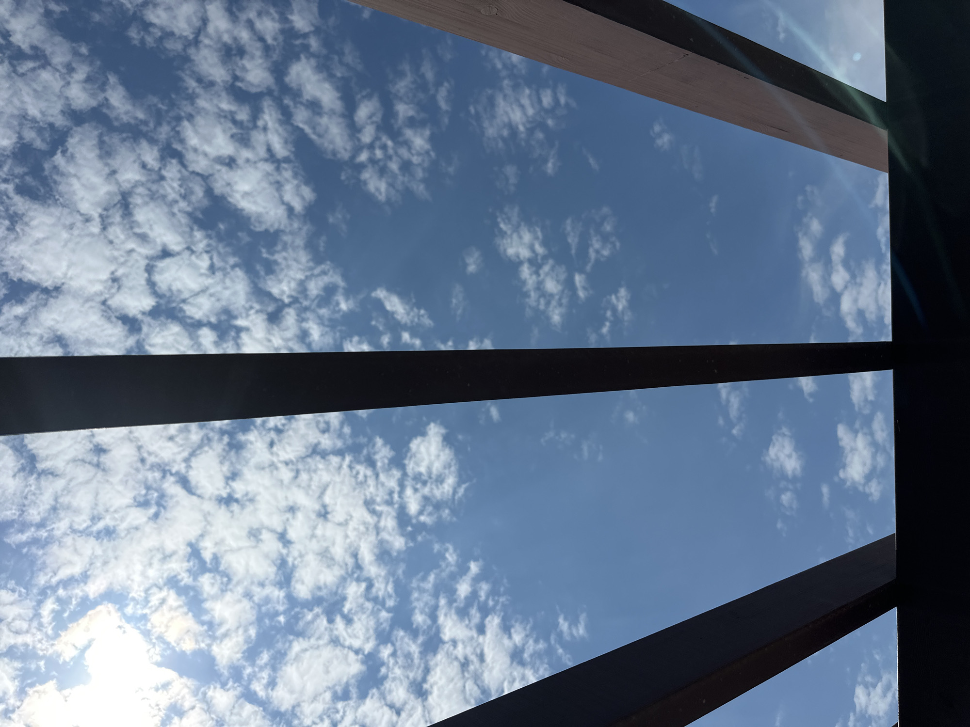 The image captures an upward perspective of a bright blue sky, generously adorned with numerous white, fluffy clouds that appear to be a mix of cumulus and stratocumulus types. The sky is partially obscured and framed by several dark, linear structural elements, likely wooden or metallic beams, which cut across the frame at various angles. These elements suggest a view from underneath a pergola, a skylight, or an open architectural roof.
Direct sunlight is visible as a strong glare and subtle lens flares in the upper right and lower left sections of the image, indicating a sunny day. The time of day appears to be during daylight hours, possibly late morning or early afternoon, given the intensity of the light and the clear visibility of the clouds against the blue sky. The weather is sunny with scattered clouds, conveying a pleasant atmosphere.
No people or specific activities are depicted in the frame. The scene focuses on the natural elements of the sky and clouds, contrasted with man-made architectural lines. There is no visible text. The photograph offers a serene and open view, taken in Trap, Montenegro.