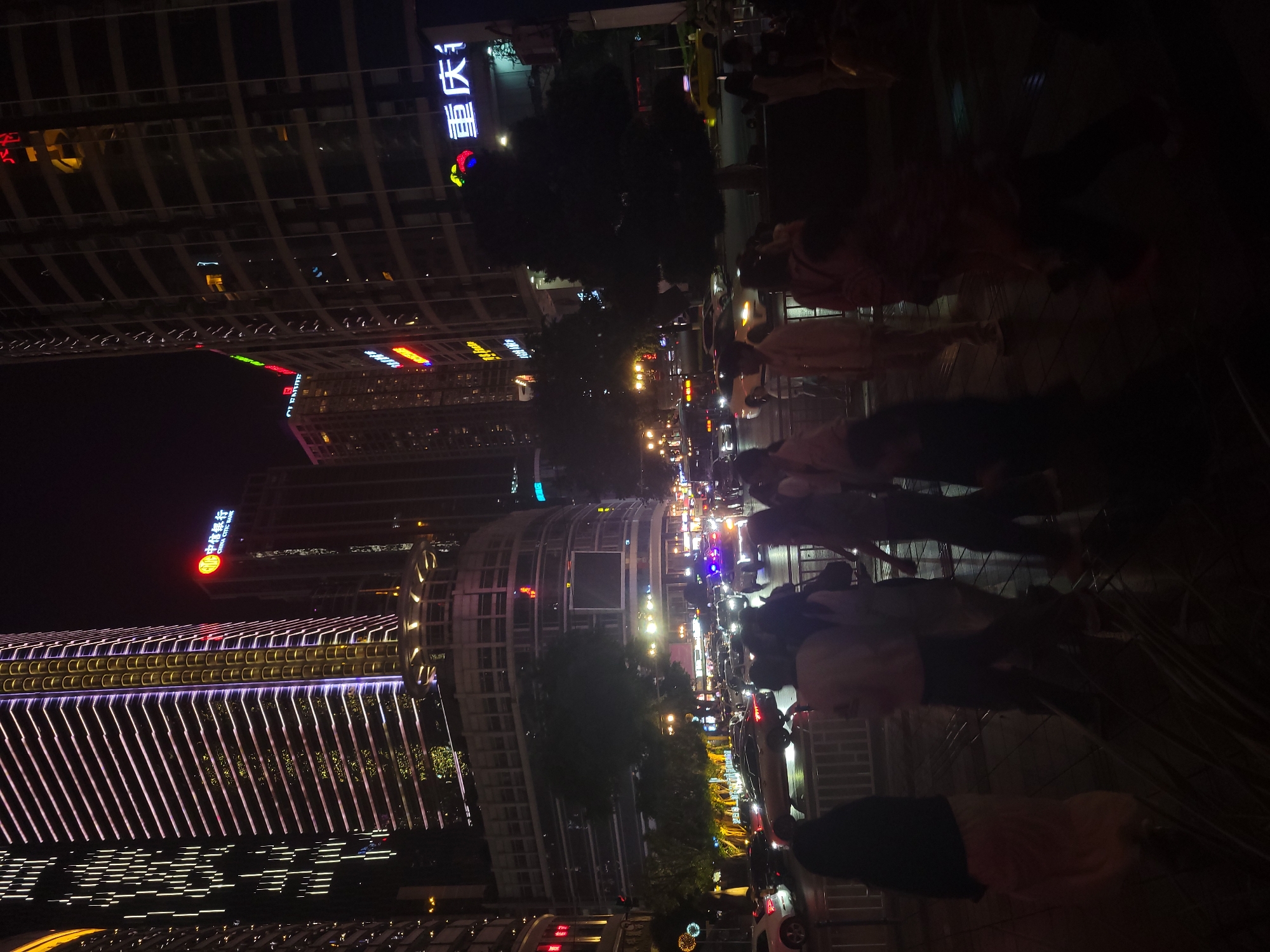 This image shows a night scene in Yuzhong District, Chongqing, China. The photograph is oriented vertically. A street runs between tall buildings with illuminated signs and decorative lighting. People walk along the sidewalk on the right. Cars and buses move along the road. Trees are visible lining the street. The buildings are modern high-rises with lit windows and neon signs in Chinese characters.