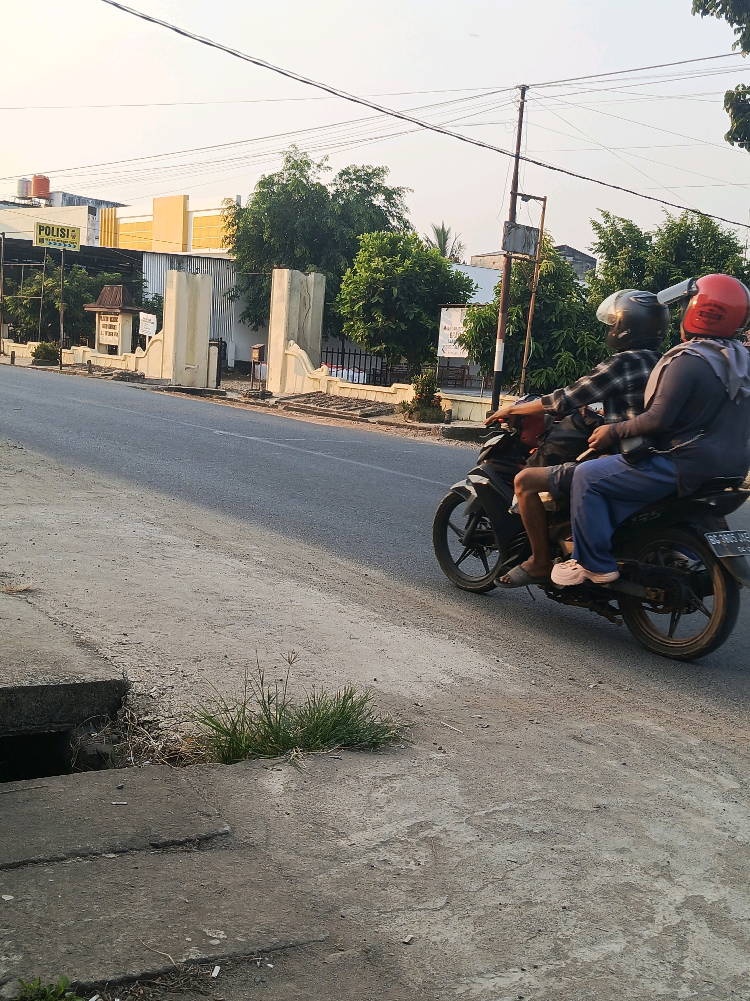 Two individuals are riding a motorcycle on a paved road in Talang Ubi, Indonesia. The person in the front is wearing a black helmet and a plaid shirt, while the person in the back is wearing a red helmet and a gray hoodie over blue pants. The motorcycle is black and has a license plate that is partially visible. In the background, there is a yellow and white building with a sign that reads "POLISI." There are also trees, power lines, and a concrete drain on the side of the road. The sky is clear and appears to be late afternoon or early evening.