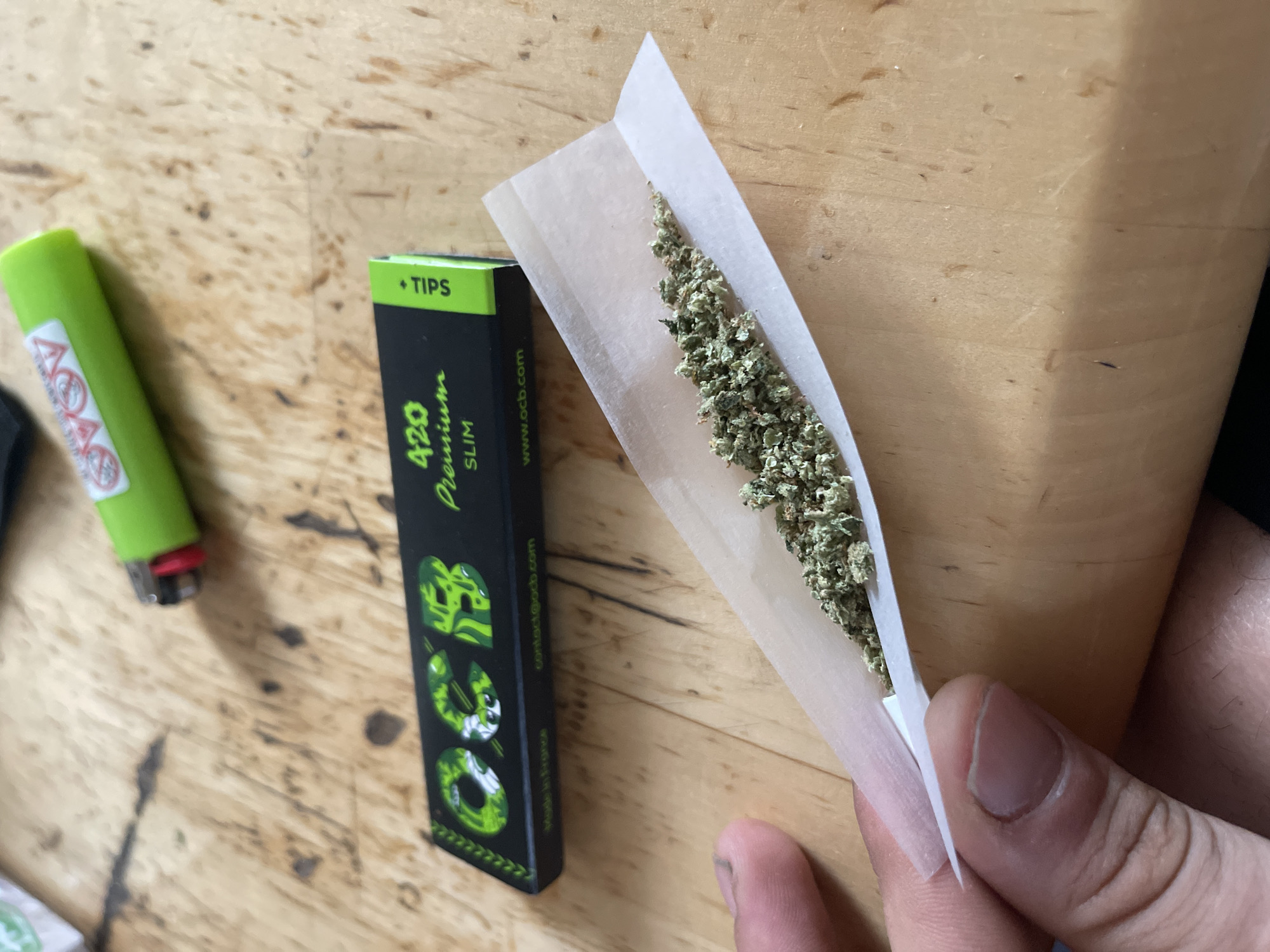 An overhead shot shows a person's left hand holding a rolling paper with cannabis on it, preparing to roll a joint. Next to the rolling paper is a pack of OCB 420 Premium Slim rolling papers and tips. A green lighter is positioned above the rolling papers. The scene is set on a wooden surface. The background is out of focus.