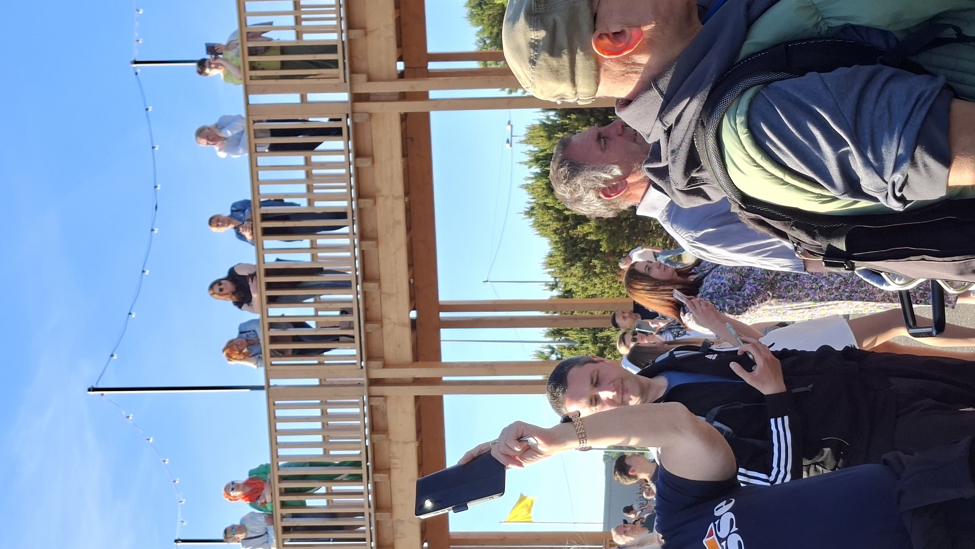 In Tallinn, Estonia, a gathering of people is observed in an outdoor setting, likely a festival or event, with a wooden structure providing elevated viewing access for some. The image captures a crowd on a sunny day, with a clear blue sky overhead.
In the foreground, a woman's arm is raised, holding a dark-colored mobile phone or tablet. To her right, a man is looking towards his left, holding a similar device. Further back, a woman in a floral dress is looking at her phone. The people on the elevated wooden walkway, in the midground, are diverse in age and attire, leaning on the railing and observing the scene below. They include individuals with red hair, dark hair, and blonde hair, some in casual clothing like jeans and t-shirts, others in more formal wear.
The environment includes trees visible in the background, and streetlights with bulbs strung across them suggest an evening event or a decorative lighting installation. The wooden structure itself is made of light-colored wood and features a railing. The overall atmosphere appears to be relaxed and social, with many individuals engaged with their mobile devices. The presence of a yellow flag and distant structures hints at a public park or event space.