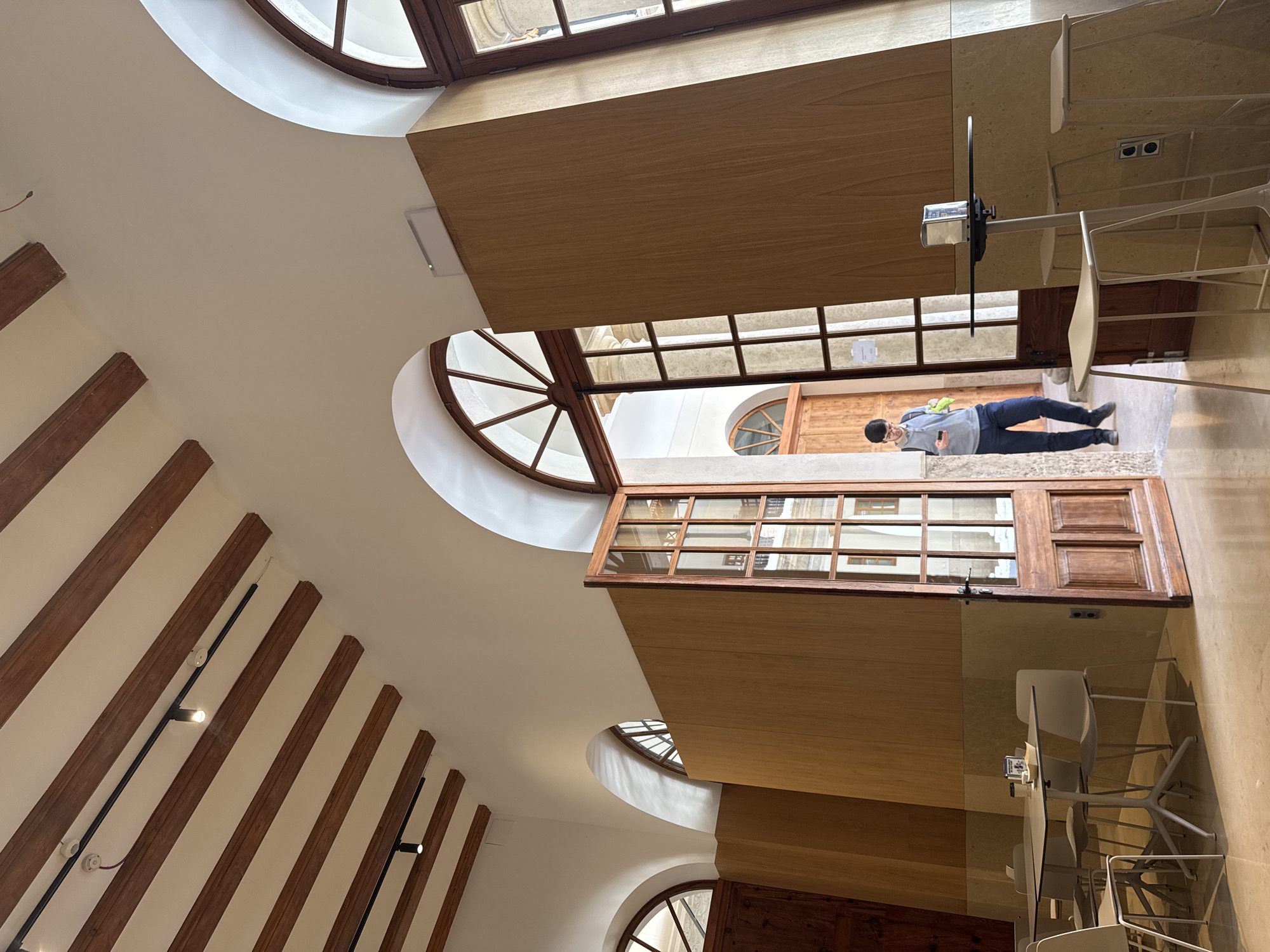 An interior shot shows an atrium in the El Carmen Cultural Centre in València, Spain. The view looks toward the ceiling, where a series of wooden beams run parallel to each other on the left side of the frame. The walls are white, and round, arched windows are visible. Wood-paneled structures and a doorway stand in the center, leading to an outdoor courtyard where a person stands. The person wears a gray sweatshirt, dark pants, and glasses and holds what appears to be a cell phone. Tables and white chairs stand to the right of the doorway.