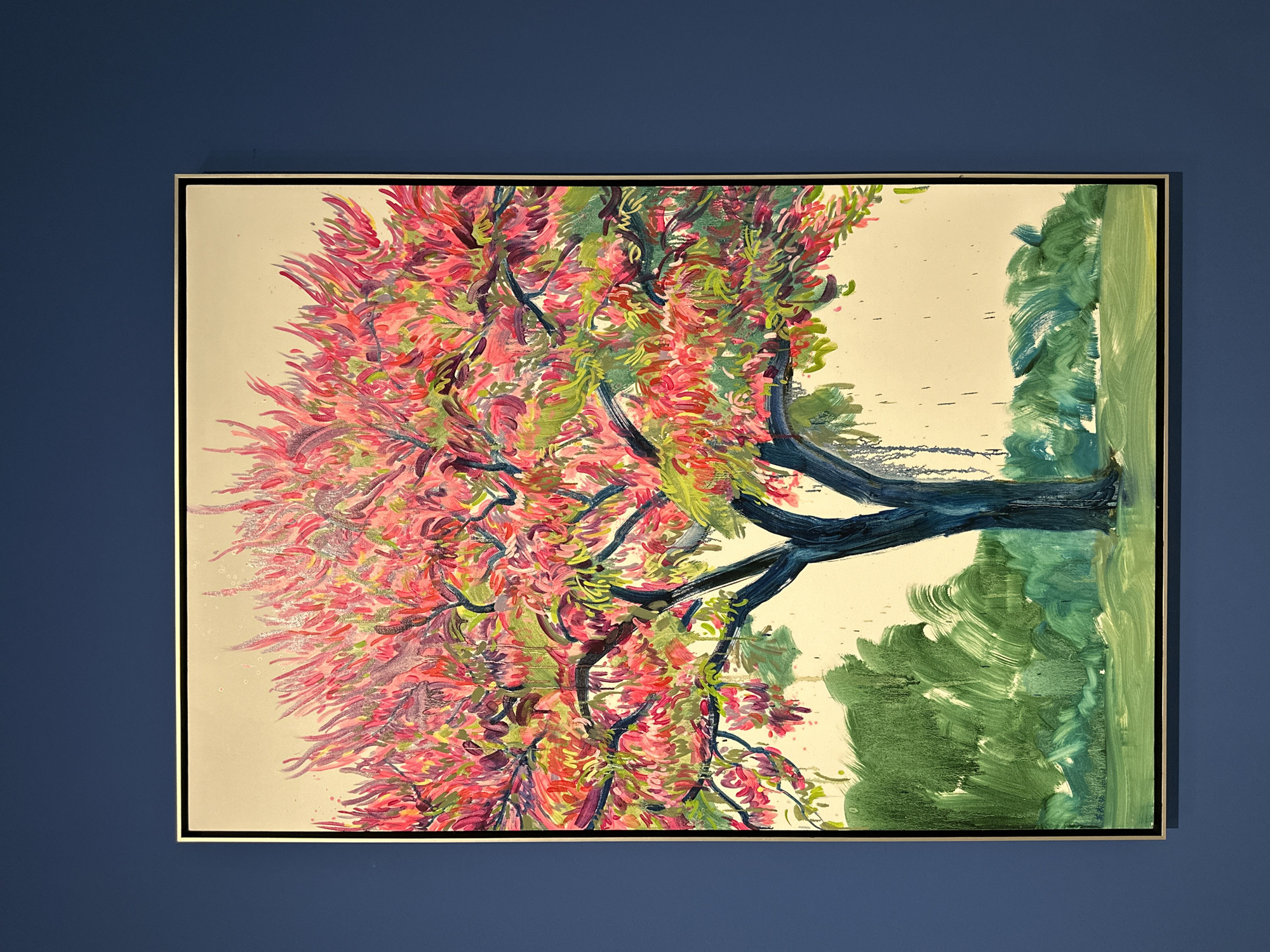 A framed painting depicting a tree with pink and green foliage is mounted on a dark blue wall. The tree's trunk and branches are rendered in shades of blue and black. The tree is positioned to the left of the frame, with a suggestion of green landscape on the right. The painting is in a gold-colored frame. The background of the painting is a light beige or cream color. There is no indication of it being in Banan, China.