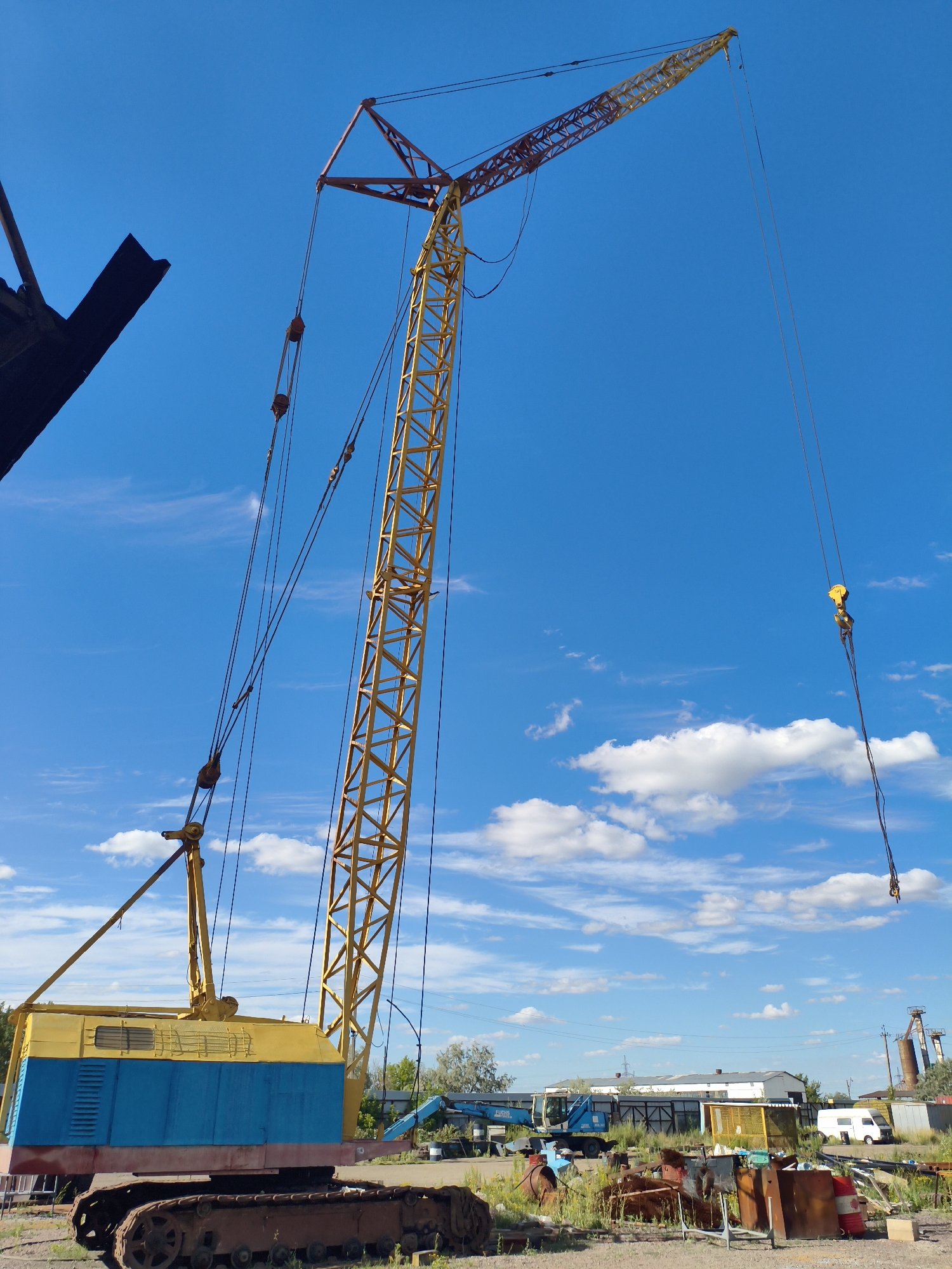 A bright yellow and blue crawler crane stands prominently against a clear blue sky dotted with white clouds. The crane's boom, a lattice structure of yellow metal, extends upwards and to the right, supporting cables and a yellow hook. The crane's base is a rusted, tracked vehicle, with a yellow and blue cabin on top. The scene appears to be an industrial or storage yard, with other assorted metal objects and structures visible in the background. A white van and some industrial buildings can be seen further in the distance. The overall impression is one of an outdoor, possibly decommissioned, construction or industrial equipment site under daylight conditions.