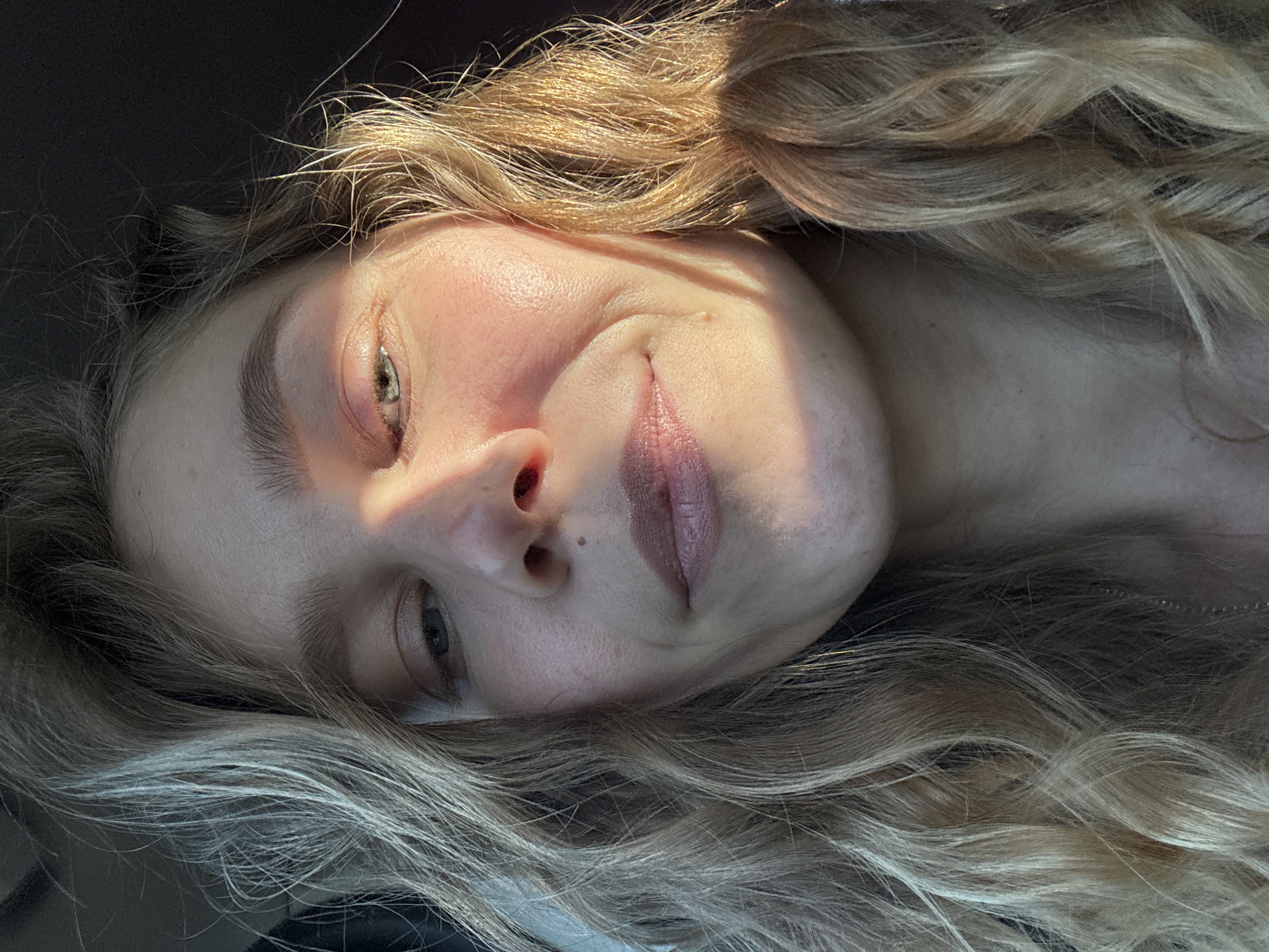 A close-up, low-angle shot depicts a woman with blonde, curly hair lying down. The sun is casting light across her face, emphasizing her right cheek, nose, and brow bone, while leaving the left side of her face partially in shadow. She has fair skin, visible pores, and a mole near her nostril. Her eyes appear light green or blue. Her lips are a muted pink. The background is dark and undefined. The location, Ново-Никольское, Russia, cannot be verified from the image alone.