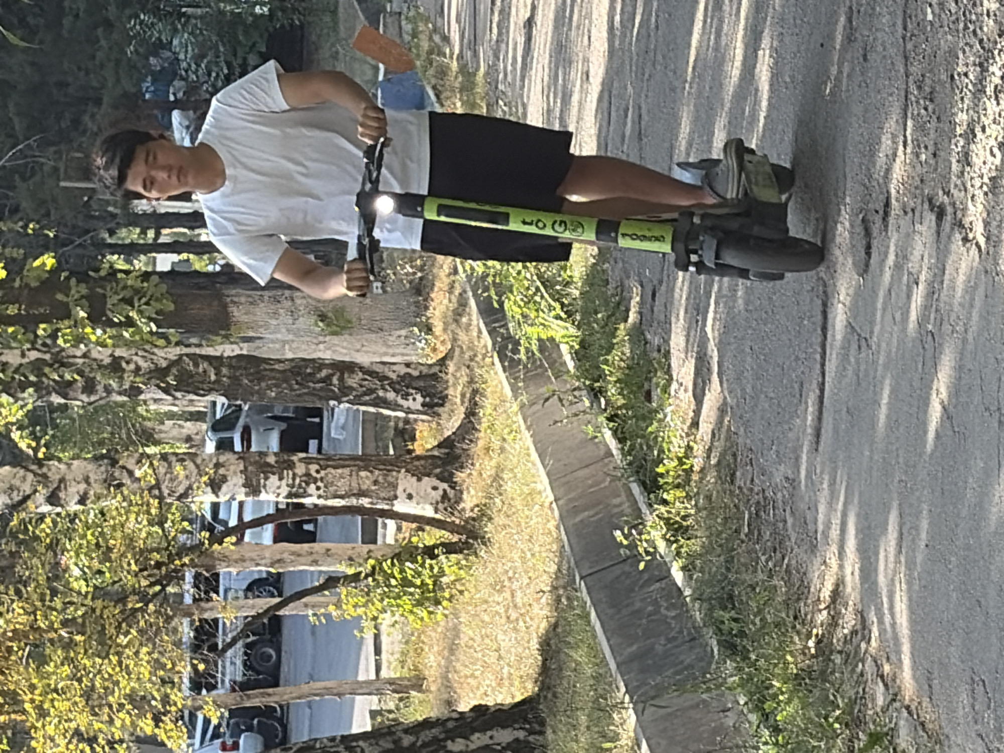 An outdoor, eye-level shot shows a young Asian man riding a green electric scooter on a paved path lined with grass and trees. The man is wearing a white t-shirt and black shorts. He is facing forward, looking towards the viewer. The scooter has a visible brand logo on the stem. In the background, several cars are parked, and trees with white bark and green foliage are prominent. Sunlight filters through the leaves, casting shadows on the path. A concrete curb runs along the left side of the path.