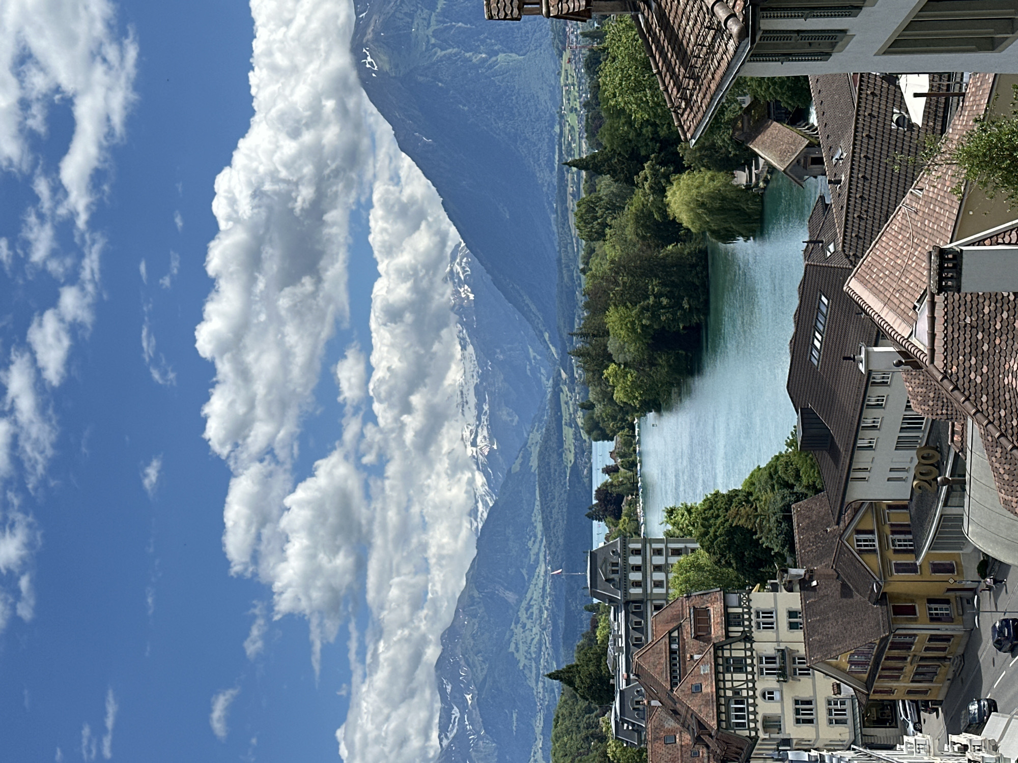This is an eye-level shot overlooking a body of water, likely a lake or river, in Thun, Switzerland. The water is a vibrant turquoise color and is bordered by lush green trees and vegetation. In the foreground, there are several buildings with red-tiled roofs, characteristic of European architecture. One building on the left has a striking half-timbered facade. Further back, a grand, multi-story building with a dark roof and many windows stands prominently near the water's edge. Beyond this, a wide expanse of water leads towards a range of majestic mountains with snow-capped peaks under a bright blue sky filled with fluffy white clouds. A small Swiss flag is visible on top of the grand building. The scene appears to be a scenic view of the Alps from Thun, with the city's rooftops and a prominent building framing the natural landscape.