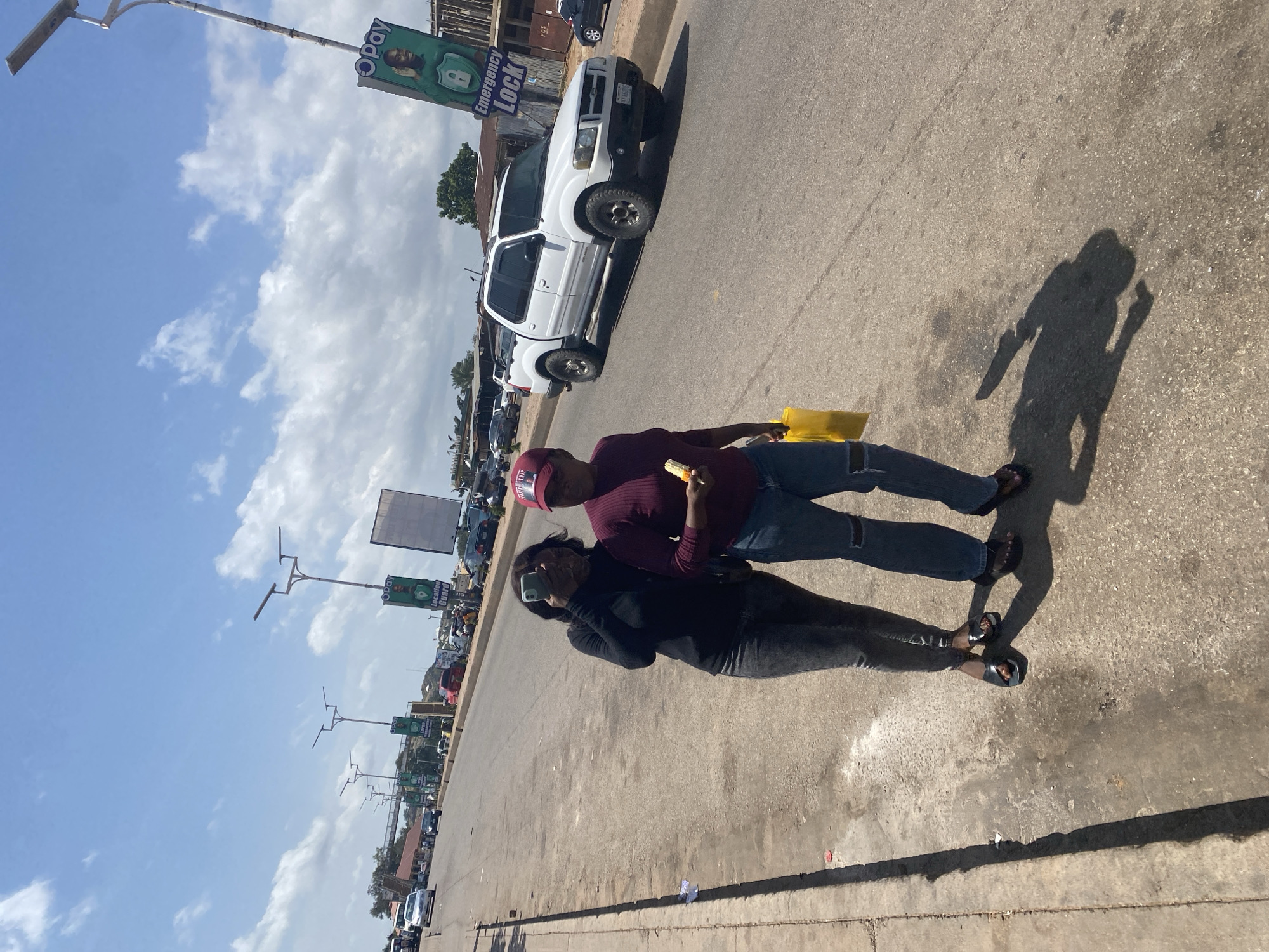 Two individuals are walking on a street in Gura Top, Nigeria. The person on the left is wearing a black long-sleeved shirt and grey ripped jeans, with black sandals. They are holding a mobile phone to their face, possibly taking a picture or video. The person on the right is wearing a maroon long-sleeved top and ripped blue jeans, with dark sandals. They are holding a yellow shopping bag in their left hand and a piece of corn in their right hand.
The street is paved and appears to be a main road with traffic. There are several vehicles visible, including a white Ford Explorer SUV parked on the right side of the road. Streetlights are present along the roadside, and there are advertisements or signs mounted on poles. The sky is blue with scattered clouds. The environment suggests an urban or semi-urban setting with buildings and trees visible in the background.