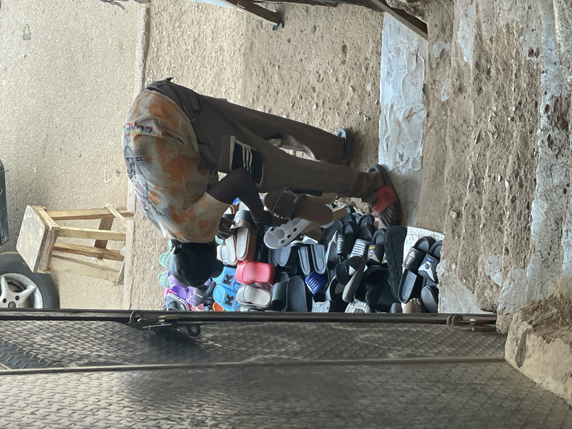 The image depicts a man in Bukuru, Nigeria, bent over selecting a pair of sandals from a large pile displayed on the ground. The man is wearing grey pants with Adidas stripes, a tie-dye shirt, and red sandals. He is positioned between a wall on the right and a vehicle on the left, with a small wooden stool also visible on the left. The backdrop suggests an outdoor sales area.