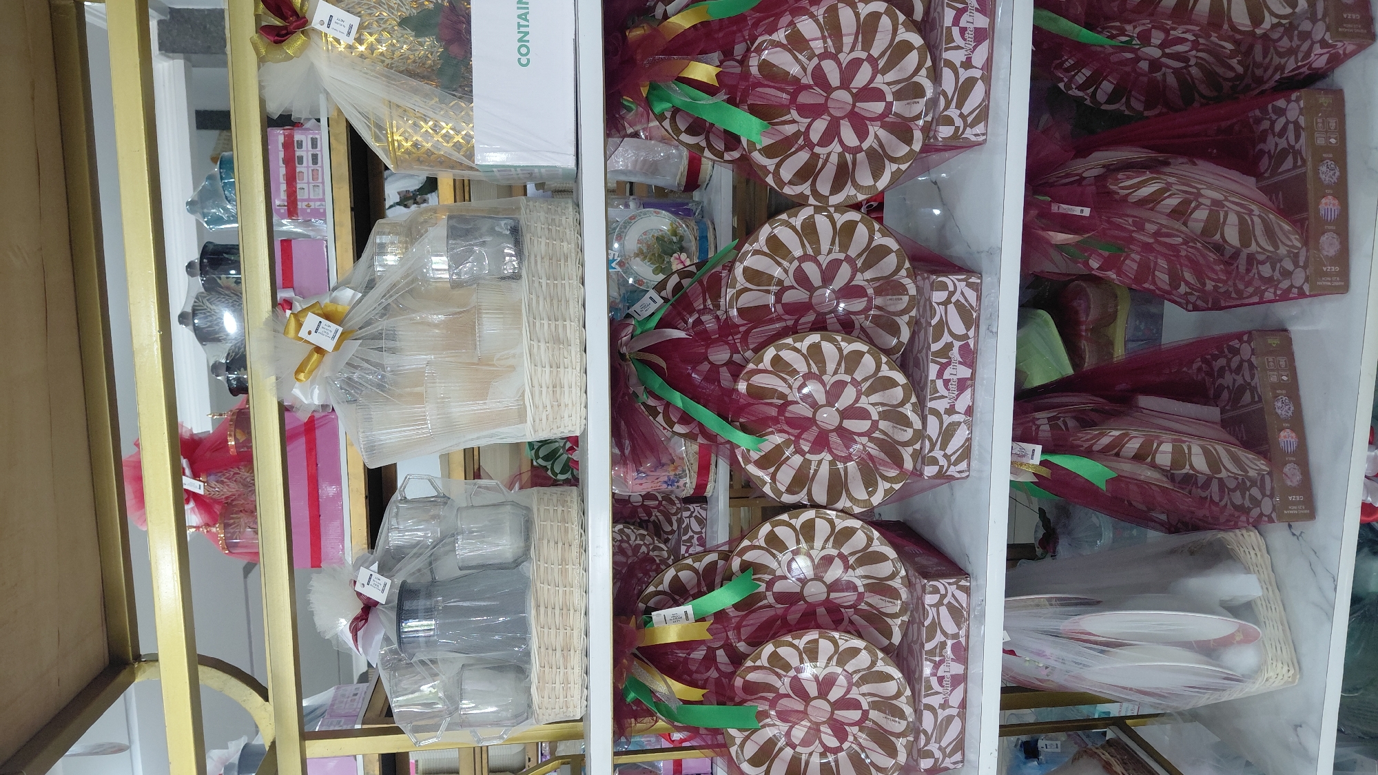 The image shows shelves in a store, possibly a gift shop or housewares store. The shelves are stocked with various items, many of which appear to be kitchenware or serving dishes, presented in gift baskets.
On the top shelf, there are several decorative items, including some that look like jars or canisters, with red and gold accents. There are also pink gift boxes.
The middle shelf features several large gift baskets. One contains a pitcher and glasses. Another appears to hold more glasses and possibly bowls. To the right, there's a golden-colored decorative item and a white box labeled "CONTAIN".
The bottom shelf displays more gift baskets. Several are covered in burgundy mesh and contain flat, round items with a floral pattern in shades of brown and white. These items appear to be plates or serving platters. Some of these baskets are placed on top of patterned boxes. To the far left, another basket contains white plates and possibly cups.
The overall impression is of a retail environment with products neatly arranged for display and sale. The lighting suggests an indoor setting. The specific location in Purwokerto, Indonesia is not visually discernible from the image itself; this detail would require external information.