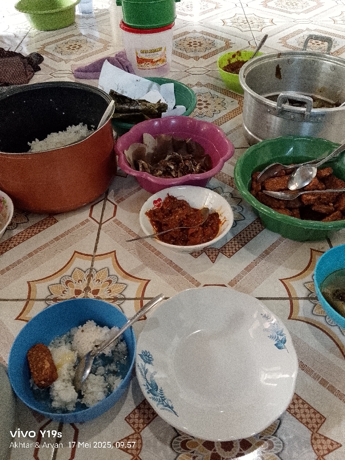 The image shows a table set for a meal in an Indonesian home in Gegesik, located at coordinates -6.538 latitude and 108.43 longitude. The table is covered with a brown and cream tiled surface. There are various dishes on the table, including a pot of white rice, a bowl of fried fish, a bowl of red chili paste, a bowl of fried tempeh, and a bowl of white rice with tempeh. There is also a large empty plate and a smaller bowl that contains white rice with tempeh. There is a spoon on the table next to the smaller bowl. In the upper left corner of the image is a small bowl with some leafy green vegetables. In the upper right corner of the image are two bowls, one containing a red sauce and the other containing a darker sauce. There is also a pot of water in the background. The scene suggests a meal is about to be served.