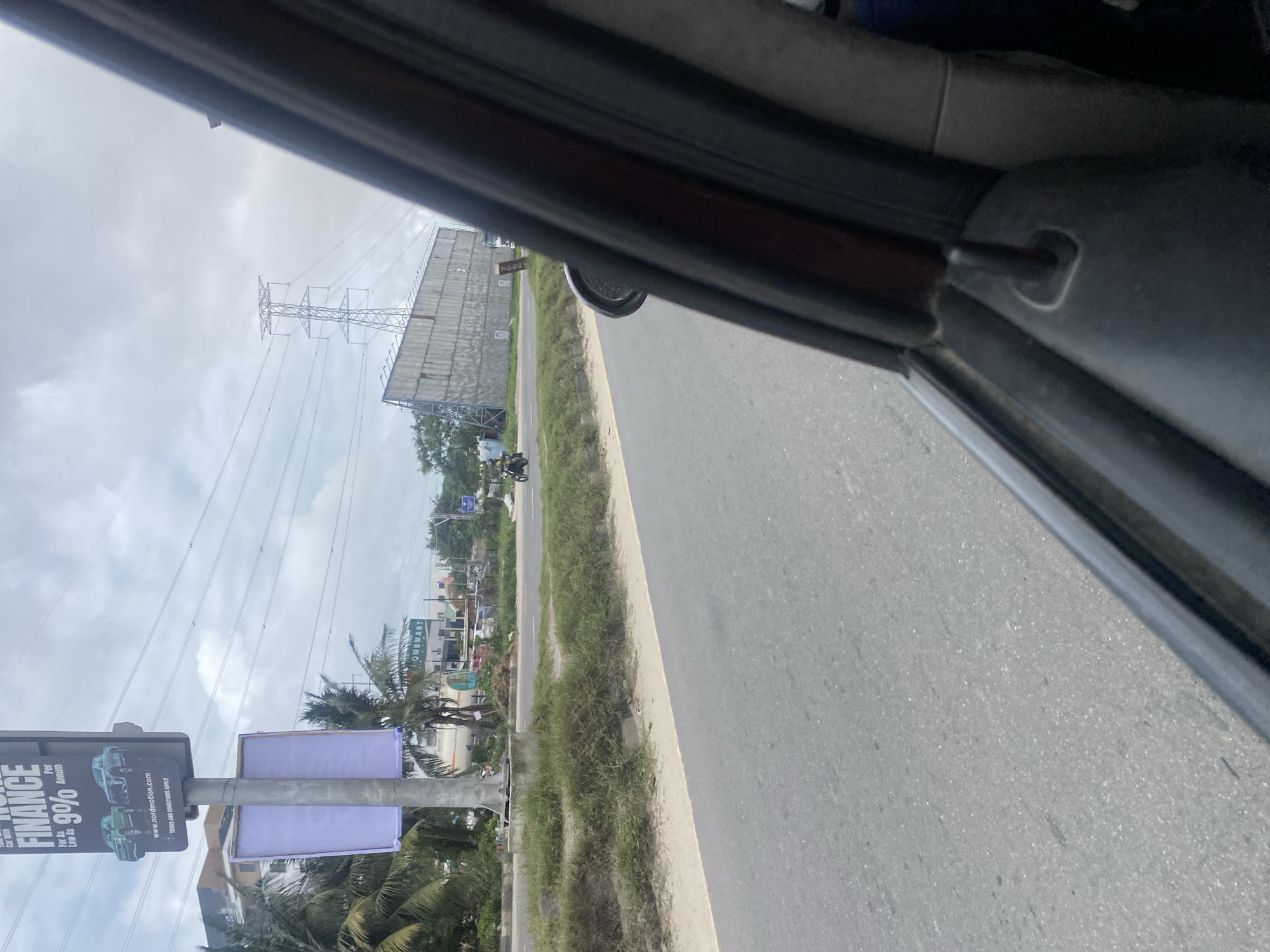 The image shows a view from inside a vehicle of a roadside scene. In the foreground, a portion of the vehicle's interior is visible, including a door frame and a side mirror. The road runs horizontally across the frame, with a grassy median strip separating it from the opposite side.
On the far side of the road, there is a large billboard with advertisements. One billboard is prominently displayed at the top of a pole, advertising "FINANCE" with a 9% annual interest rate and an image of two cars. Below this is a white, blank banner. Further down the road, a large, grey-paneled billboard stands at an angle, with power lines and a large transmission tower in the background.
Several palm trees and other vegetation are visible along the roadside. In the middle ground, a person on a motorcycle is traveling away from the viewer. In the distance, a building with signage that appears to say "MBMART" can be seen. The sky is overcast with clouds. The overall environment suggests an urban or semi-urban setting. The image is taken from a moving vehicle, resulting in a slight blur.