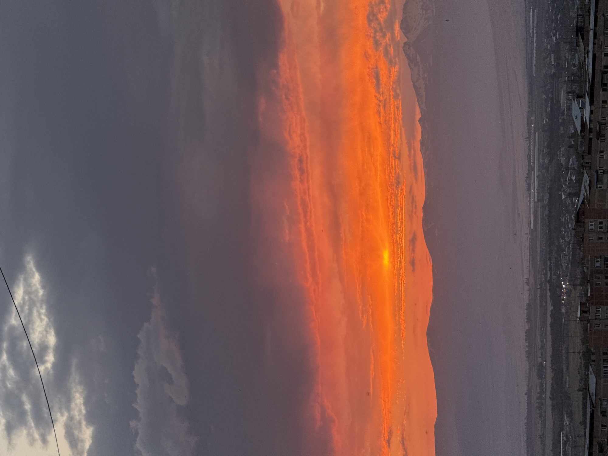 The image depicts a vibrant sunset over the city of Abovyan, Armenia. The sky is ablaze with intense oranges and reds, with streaks of pink and purple, indicating a dramatic atmospheric phenomenon. Dark, heavy clouds gather in the upper portion of the sky, contrasting with the fiery hues below. A faint wire stretches across the top left of the frame.
Below the captivating sky, a range of mountains forms a majestic backdrop. The peaks of these mountains are dusted with snow, creating a stark contrast with the warm colors of the sunset. The middle ground features a hazy, purplish landscape, hinting at the distance between the viewer and the mountains, possibly containing fields or lower hills.
The foreground of the image reveals a detailed view of the city of Abovyan. Numerous buildings, characterized by their flat roofs and concrete structures, are visible. Some structures appear to be apartment buildings with visible windows, while others may be commercial or industrial buildings. Scattered lights indicate the presence of artificial illumination within the city. The urban landscape extends across the lower portion of the frame, leading the eye towards the expansive natural scenery beyond. Several small, dark specks, likely birds, are visible in the sky and over the landscape.