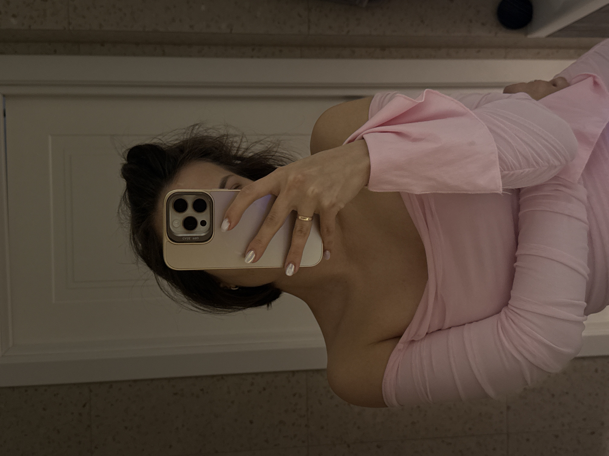 This is a mirror selfie showing a person, likely female, with dark hair tied back, posing indoors. The image is rotated 90 degrees counter-clockwise. The individual is wearing a light pink, long-sleeved top, possibly an off-the-shoulder style, with a textured or ribbed fabric visible on the sleeves. Her face is largely obscured by a light purple/lavender smartphone, which she holds with her right hand. The phone features a triple camera lens array, and the partial word "CYBE" is visible in reverse on its case. Her fingers are painted with light, possibly iridescent, nail polish, and a simple gold-colored ring adorns her ring finger. In the background reflected in the mirror, a white architectural element, possibly a door or cabinet frame, is seen behind the person. Above this, a darker, subtly textured wall is visible, while below, a speckled or tiled surface suggests a floor or countertop. The setting appears to be an interior space, likely a bathroom or dressing room. The location context provided is Pervomaysky, Russia.