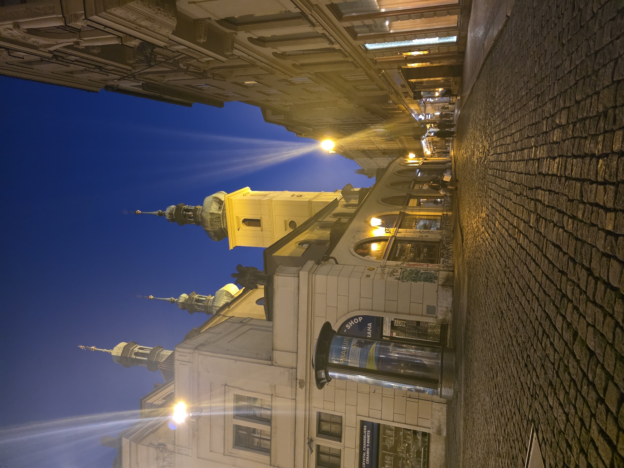 The image depicts a cobblestone street in an urban environment at dusk or night. On the left side of the street, there is a pale building with architectural features including arched windows and a prominent tower with a green dome and spire. The building has several windows and a shopfront at street level with signage that reads "PRAGUE SHOP". There is a cylindrical display pillar in front of the building. Further down the street, more buildings line the right side, featuring illuminated shop windows and upper floors with arched windows. People are visible walking along the street and sidewalks, some carrying bags. The sky is a deep blue, and dramatic beams of light emanate from a source not visible in the frame, casting rays upwards and across the scene. Streetlights are also visible, illuminating the cobblestone pavement and casting a warm glow. The overall atmosphere is one of a European city at night, with historical architecture and commercial activity.