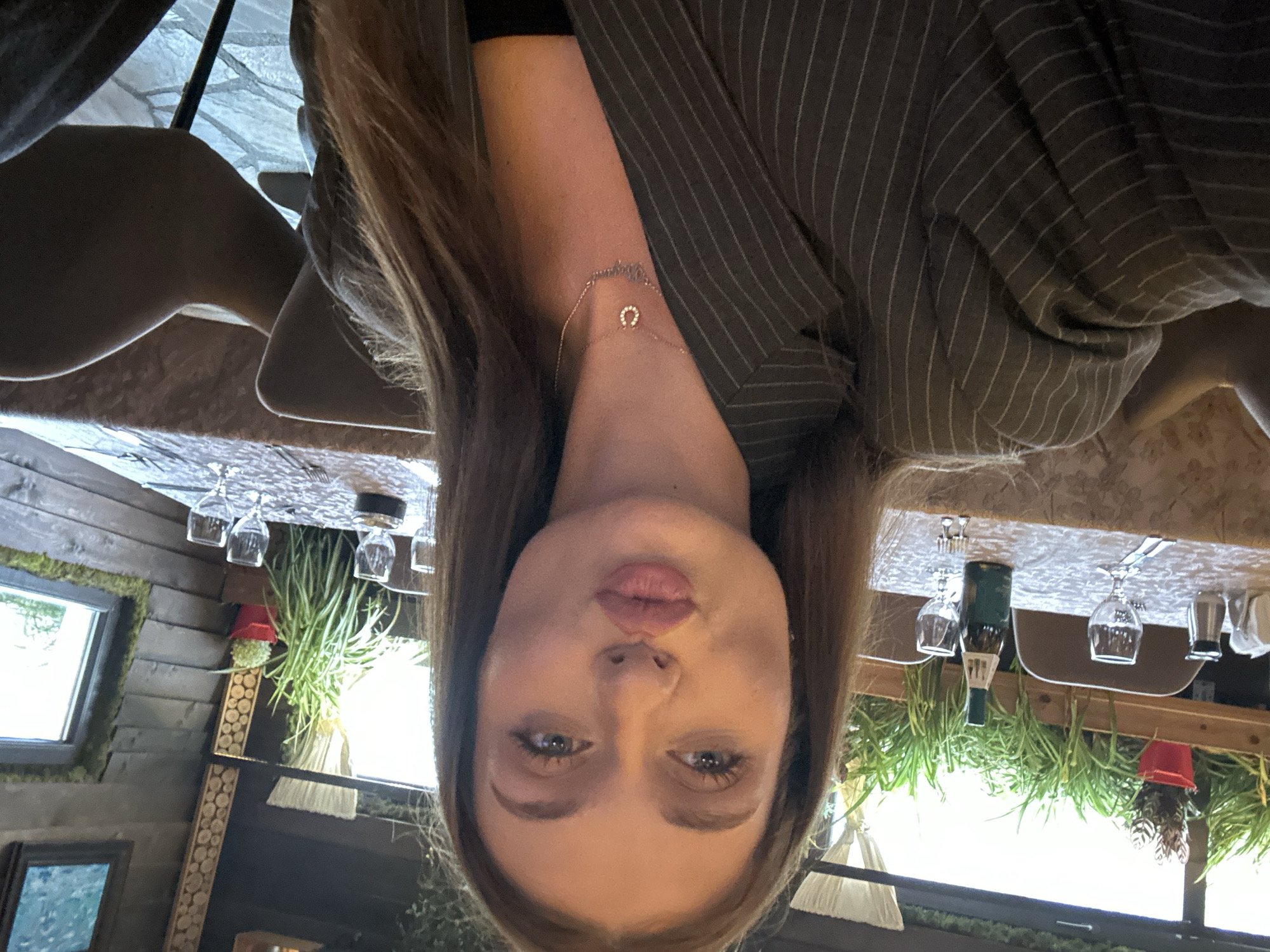 The image shows a woman with long brown hair wearing a pinstripe blazer and a delicate necklace with a horseshoe pendant. She appears to be at a restaurant. In the background, there is a wooden wall adorned with moss and framed pictures, and windows are visible. The table is set with wine glasses, silverware, and a bottle of wine. Plants are suspended from the ceiling near the windows, giving the location a botanical theme. The restaurant likely is in Sofia, Bulgaria, given the user's request.