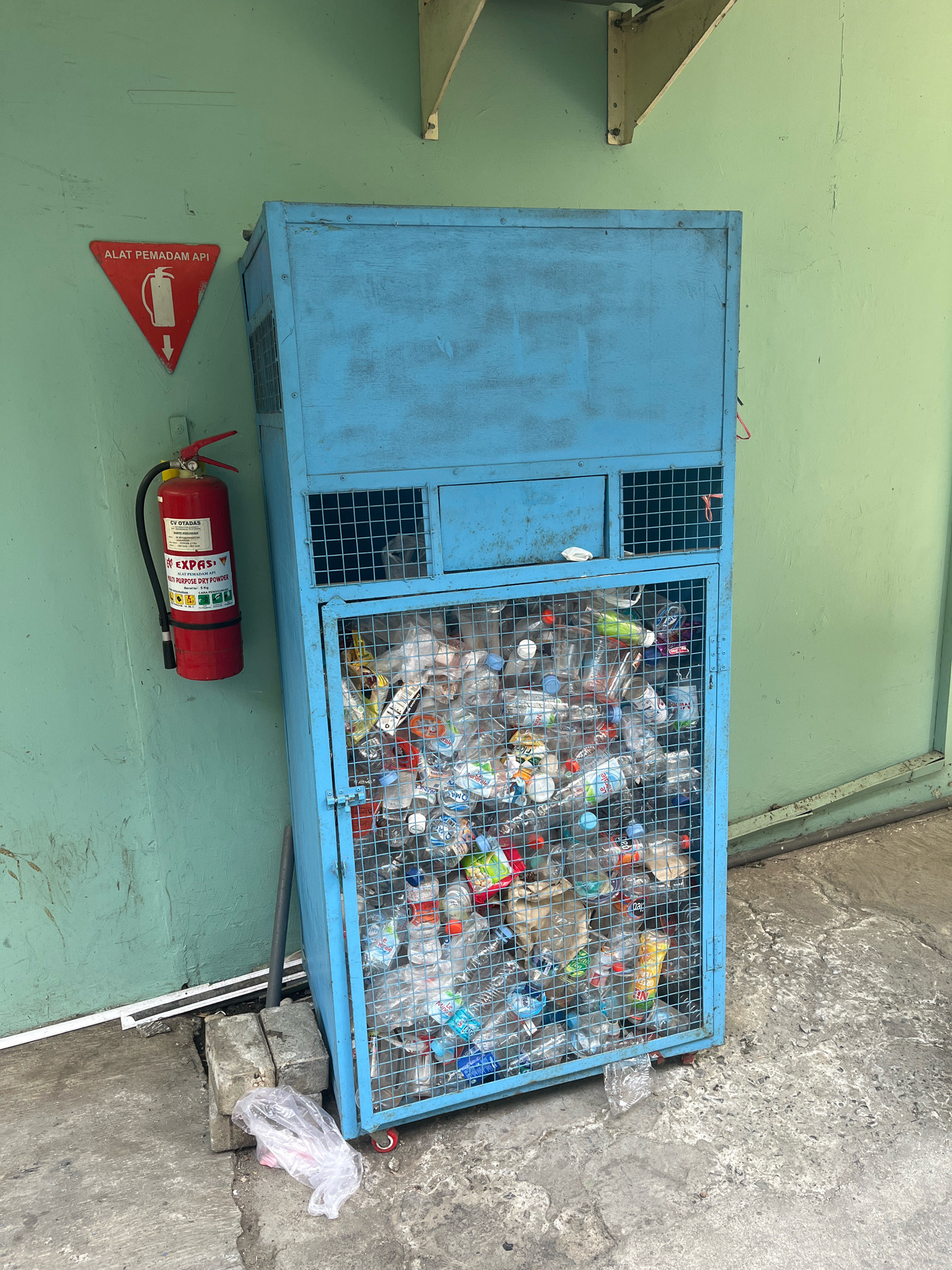 This image captures an outdoor scene in Dapur, Indonesia, featuring a blue metal recycling bin filled with plastic bottles. To the left of the bin, a red fire extinguisher is mounted on a pale green wall, accompanied by a triangular red sign with a white fire extinguisher symbol and the text "ALAT PEMADAM API." The recycling bin itself is a large, cage-like structure with a solid blue top and front panel. The lower section of the bin is open, revealing a dense collection of clear plastic bottles of various sizes and brands. The ground is concrete, with some debris including a plastic bag and concrete blocks visible near the base of the bin. The environment appears to be an outdoor service area or alleyway.