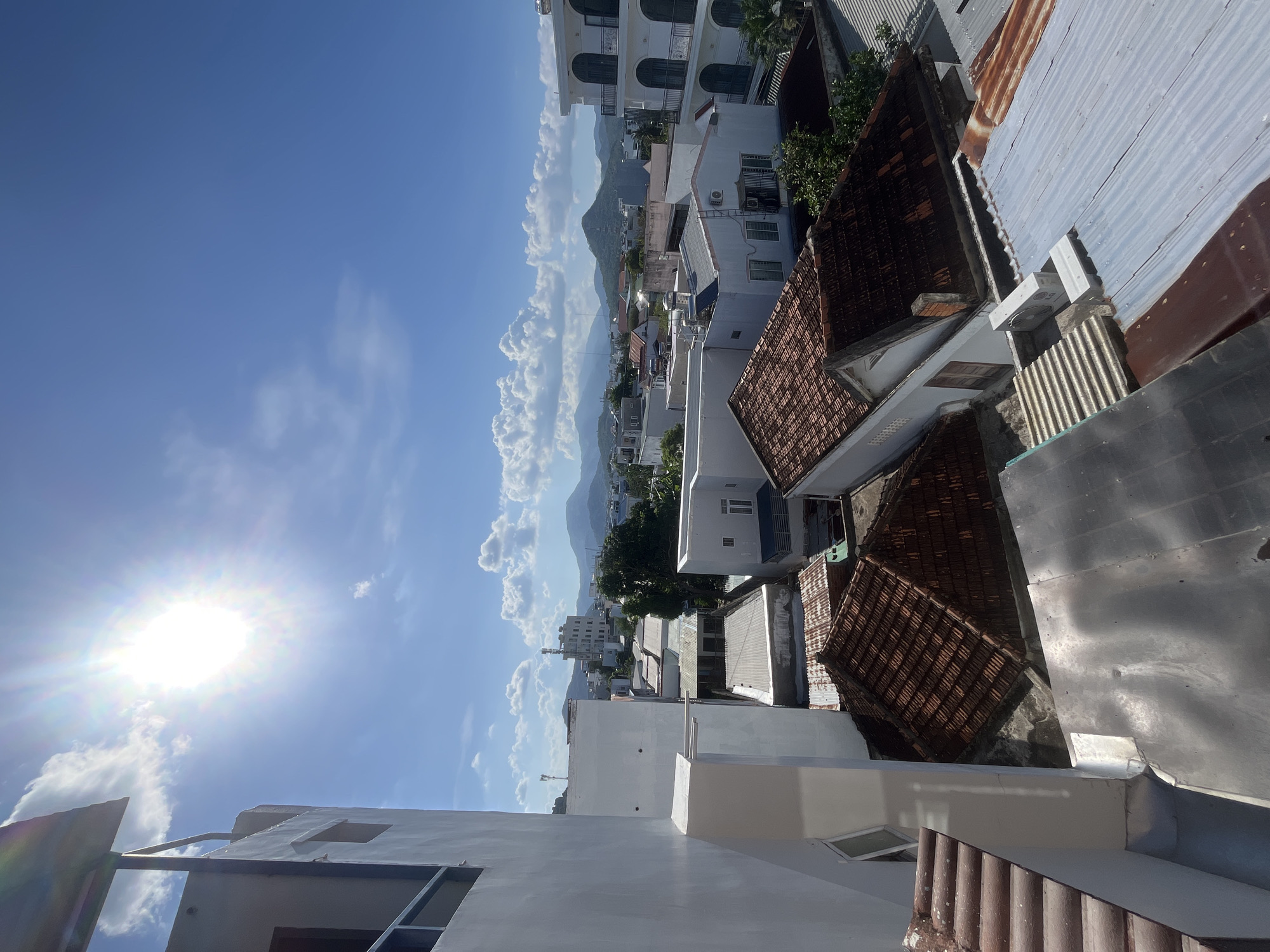 The image captures a daytime scene from an elevated perspective, presumably from the rooftop of a building in Bac Nha Trang Ward, Vietnam. The foreground consists of portions of the building's structure, including a white-painted section with a window and corrugated roofing. Beyond the foreground, there's a patchwork of rooftops, a mix of white, brown-tiled, and corrugated metal. In the distance, a mountain range forms the backdrop, partially obscured by a line of puffy white clouds extending down to the urban area. The sky is predominantly blue with scattered clouds. The sun is visible, positioned in the upper left, causing a significant glare and bright illumination.