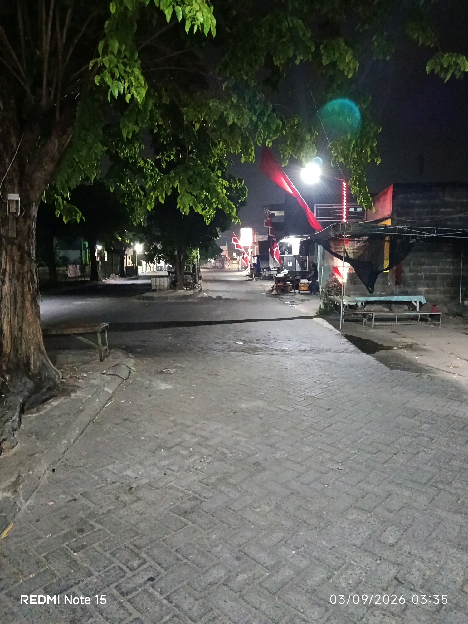 The image shows a nighttime scene in a street in Keboansikep, Indonesia. A large tree with green foliage is prominent on the left side of the frame, with a bench placed beneath it. The street is paved with interlocking bricks and appears to be a pedestrian area or a narrow road. To the right, there are several structures that appear to be stalls or small shops, some with red and white decorations. Lights illuminate the area, and a red cloth is draped from one of the structures. A person is visible standing near one of the stalls. The overall impression is of a modest, possibly commercial street at night.