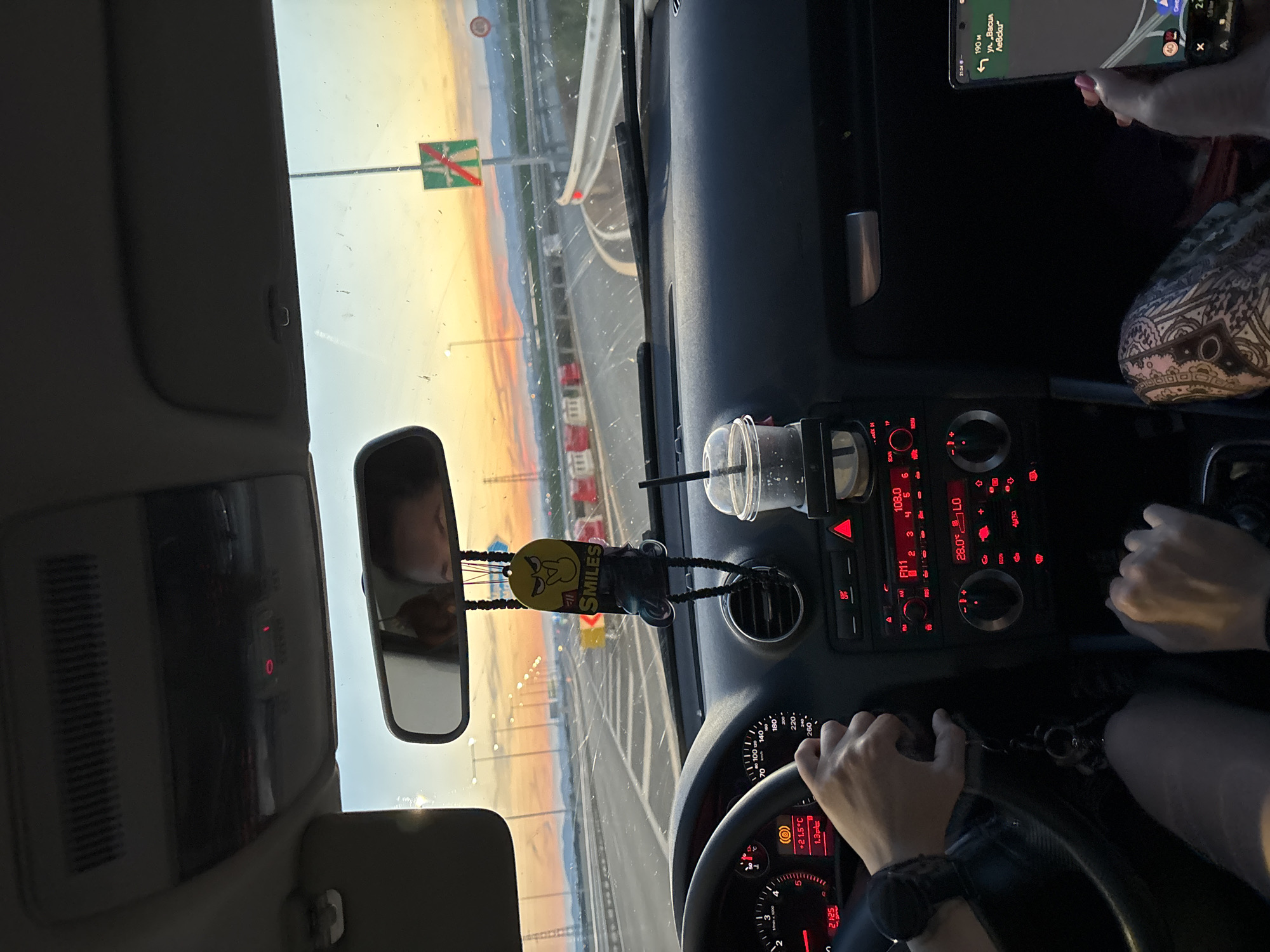 This is a point-of-view shot from inside a car traveling on a highway at sunset. The driver's hands are on the steering wheel, and the dashboard displays show speed, temperature, and radio information. A beverage cup with a straw is in the cup holder, and a car air freshener hangs from the rearview mirror. The exterior view through the windshield shows a highway with oncoming traffic, road barriers, streetlights, and a sunset sky. A road sign indicating an upcoming turn is visible. The image is likely taken in or near Mramor, Bulgaria, given the context.