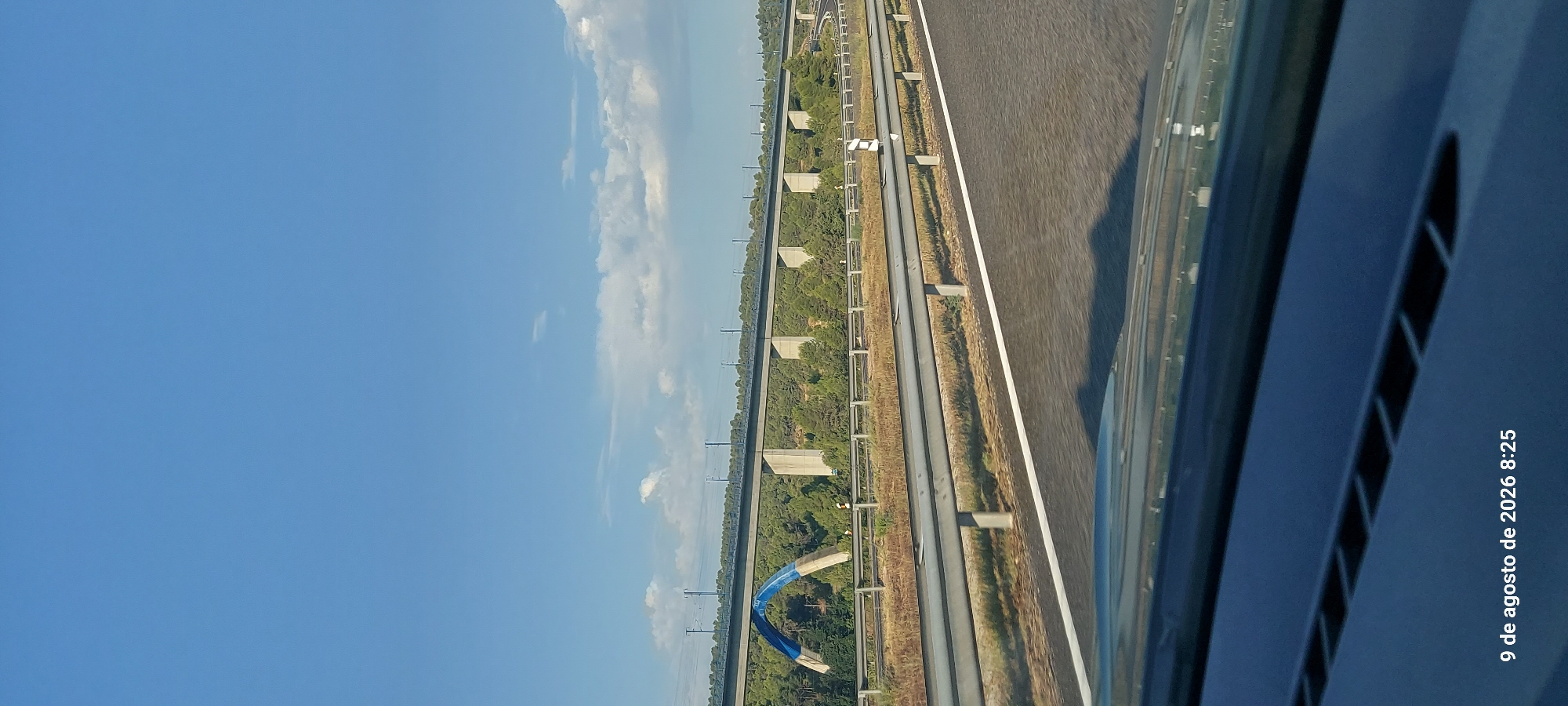 The image shows a view from a car driving on a highway in Vinaixa, Spain, on August 9, 2026, at 8:25 AM. The sky is blue with scattered clouds. In the distance, a large viaduct, designed for a high-speed railway, spans across a forested area. The viaduct is supported by numerous concrete pillars and features a striking blue archway on the left side. The foreground shows the asphalt road with white lane markings and a metal guardrail separating the highway from the shoulder. The reflection of the sky and trees can be seen on the car's dashboard in the lower part of the image. The environment appears to be dry and sunny, typical of summer in the region.