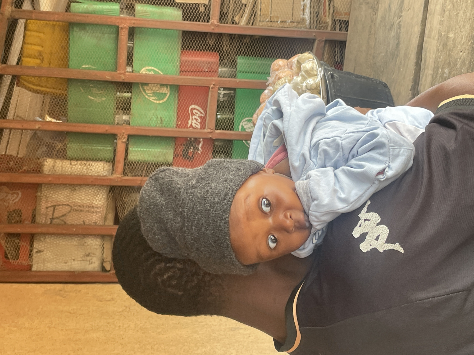 A person's back and shoulder are visible, carrying a baby. The person is wearing a black t-shirt with a white Kappa logo on the left side of the chest and a yellow and black striped trim on the collar. The baby is wearing a grey knit beanie and a light blue hooded jacket. The baby's face is visible, looking towards the left of the frame with wide eyes. The background shows a shelf or display unit filled with stacked green and red crates, some with visible branding such as "Coca-Cola" and "Pepsi". There are also yellow containers and some white bags with writing on them. A black bucket filled with what appears to be food items like plantains is also in the background. The overall setting suggests a market or shop environment. The lighting indicates it is daytime. The image captures a moment of a person carrying a child in a busy commercial area, possibly a street-side stall or small shop, which are common in areas like the Tudun Wada district of Jos, Nigeria.