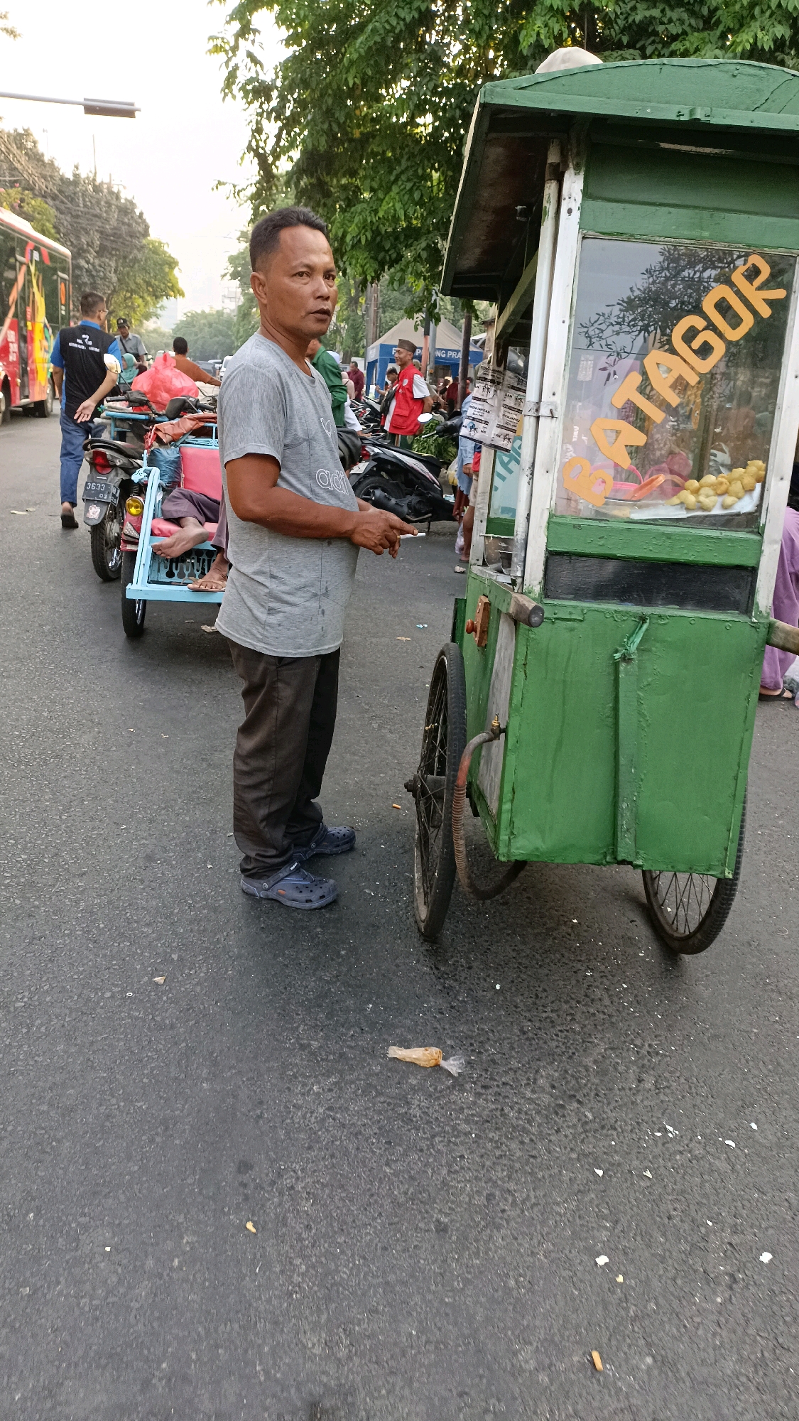 The image depicts a street scene in Surabaya, Indonesia, during daylight hours. A man, appearing to be in his late 40s or early 50s, stands beside a green, wheeled food cart. The cart has a glass display showing food items, and "BATAGOR" is prominently written in yellow lettering on the glass. The man is wearing a grey t-shirt and dark pants, with dark blue clogs on his feet. He is holding a cigarette in his right hand.
Behind the man and to his left, a motorbike is parked, with a blue seat and a person partially visible lying on it, wearing a red garment. Further to the left, a red and white bus is visible, along with other vehicles and people on the street, suggesting a busy urban environment. The ground is paved asphalt, with some scattered debris, including a plastic bag with contents on the right side of the frame. Trees and foliage are visible in the background, indicating an outdoor setting. The lighting suggests it is either early morning or late afternoon.