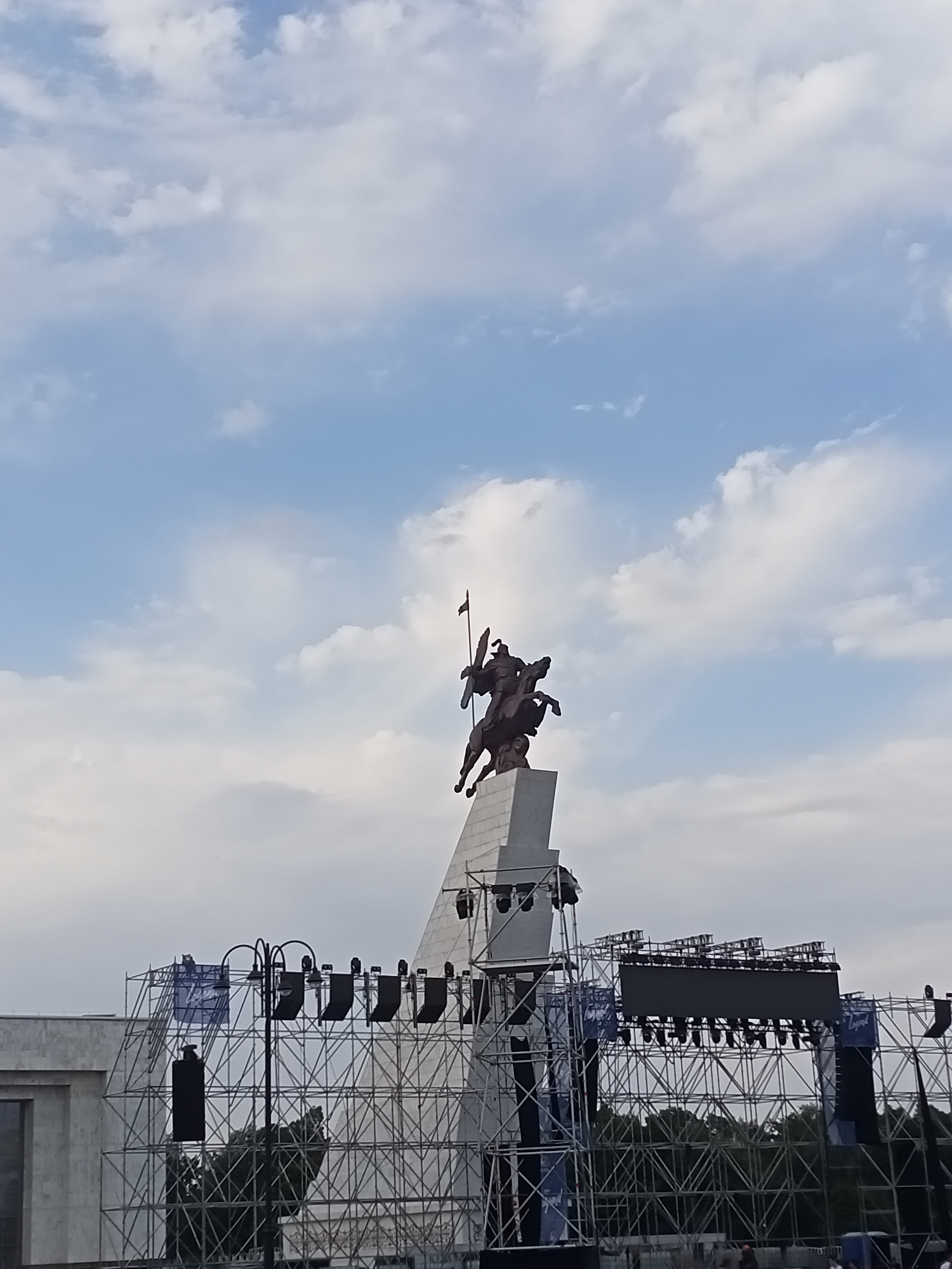 The image captures a bronze equestrian statue on a tall pedestal against a sky filled with white clouds and blue patches. The statue depicts a warrior on horseback, holding a flag. Below the statue, a large metal scaffolding structure is erected, featuring multiple tiers of lights and a large screen. Banners with blue and white designs are visible on the scaffolding. To the left of the scaffolding, a portion of a light-colored building is visible. Trees are seen behind the scaffolding. The scene appears to be set outdoors, likely in a public space like a square or park, possibly during the setup for an event or performance. This is a view of the Ala-Too Square in Bishkek, Kyrgyzstan, where the Independence Monument, an equestrian statue, is located.