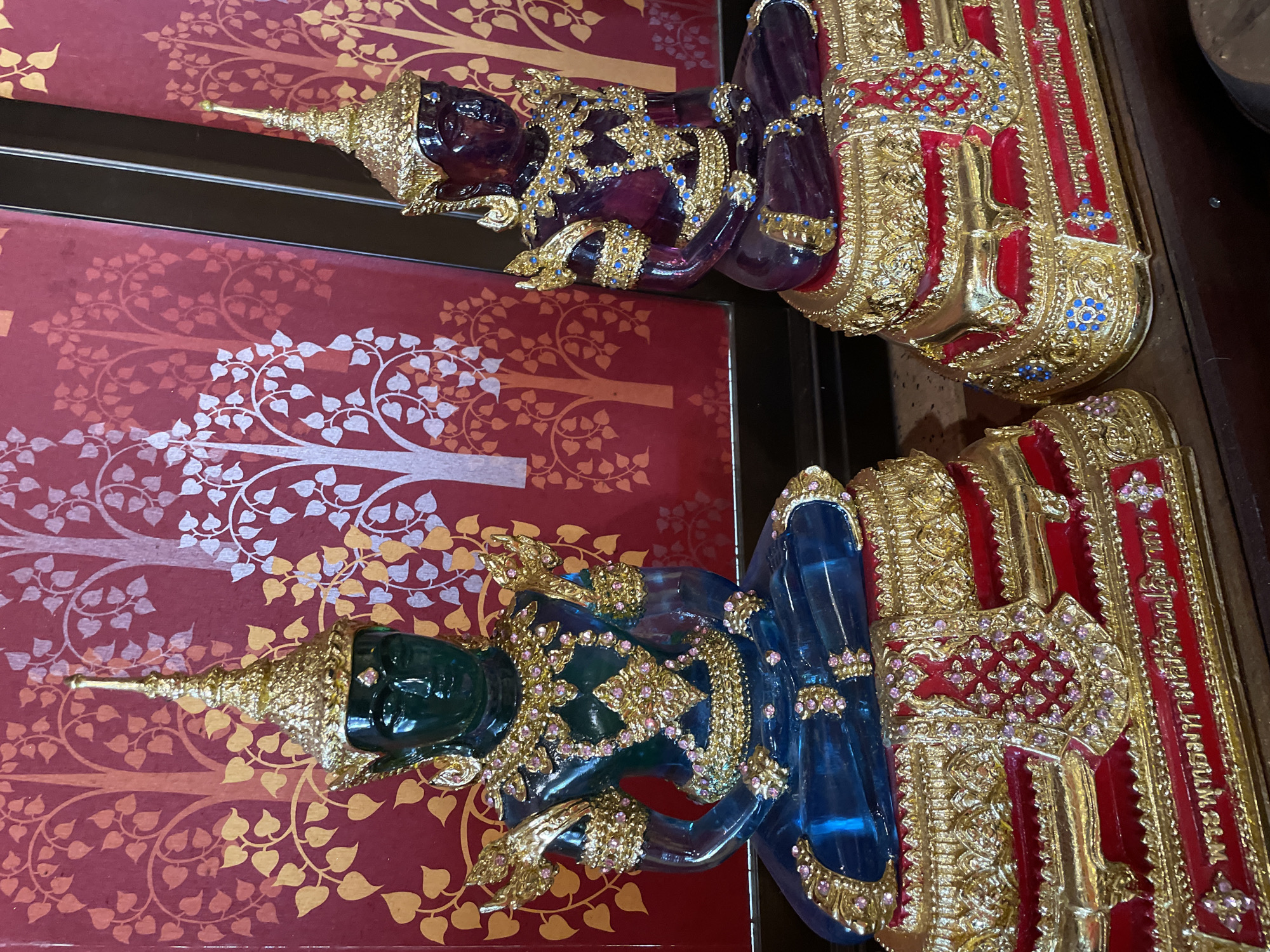 The image depicts two ornate statues of Buddha figures seated in meditation. The statue on the left is predominantly green and blue, while the one on the right is a deep purple. Both figures are adorned with intricate gold embellishments, including crowns, necklaces, armbands, and waistbands, which are further decorated with colorful small gemstones. The statues are seated on elaborate gilded bases, red in color and decorated with similar golden patterns and small blue and pink gemstones. The backdrop is a vibrant red fabric with a repeating pattern of stylized trees and leaves in gold and white. The statues are positioned on a dark wooden surface. The scene is likely a display within a shop or a religious establishment in Bang Sare, Thailand, given the cultural context of such artistic representations.